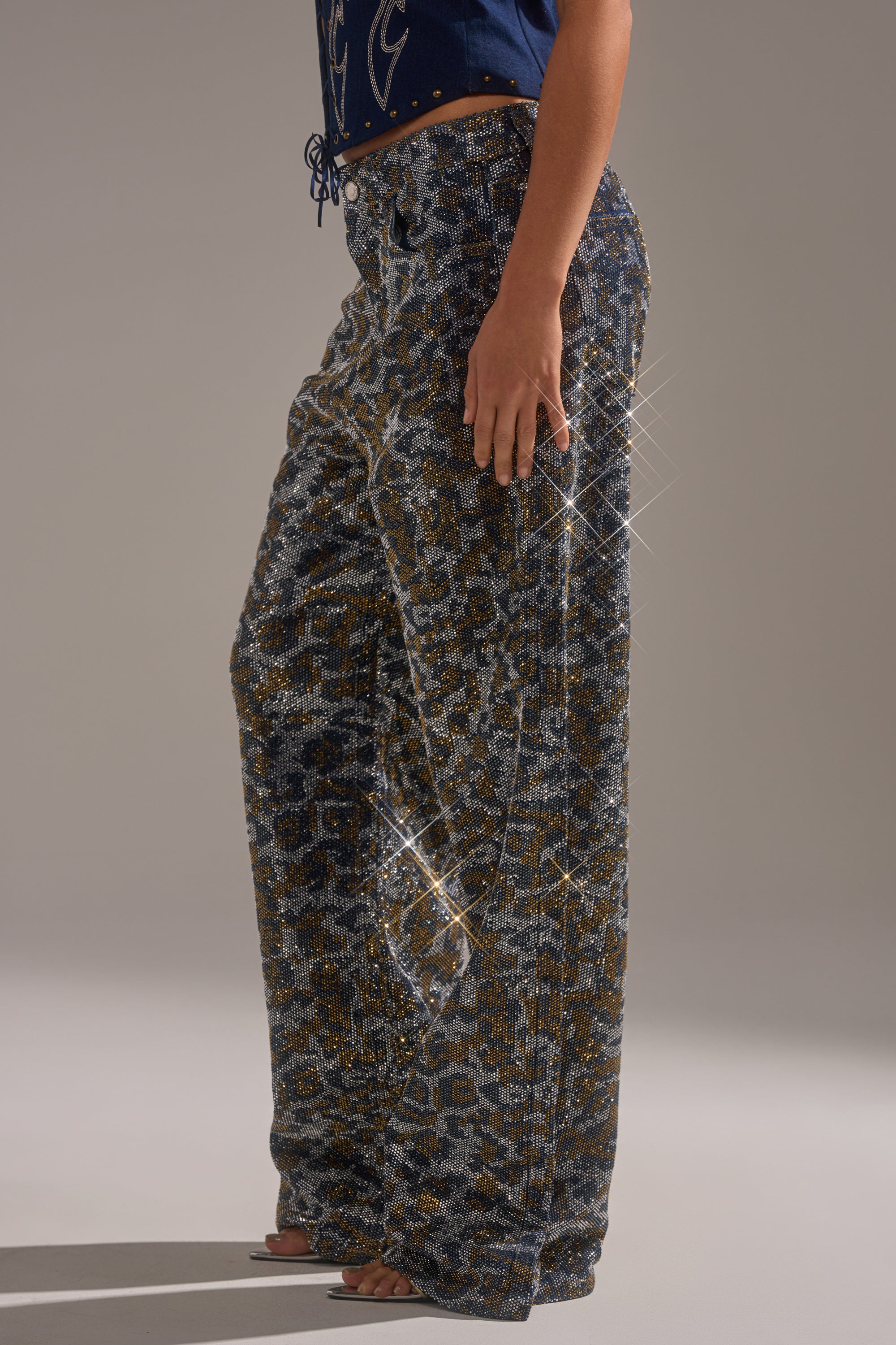 A person stands sideways, wearing the CHEETAH GIRLY RHINESTONE DENIM PANT—wide-leg jeans with glittery blue, gold, and silver leopard print. Their hand is on their hip and the hem covers their feet against a neutral background. 2