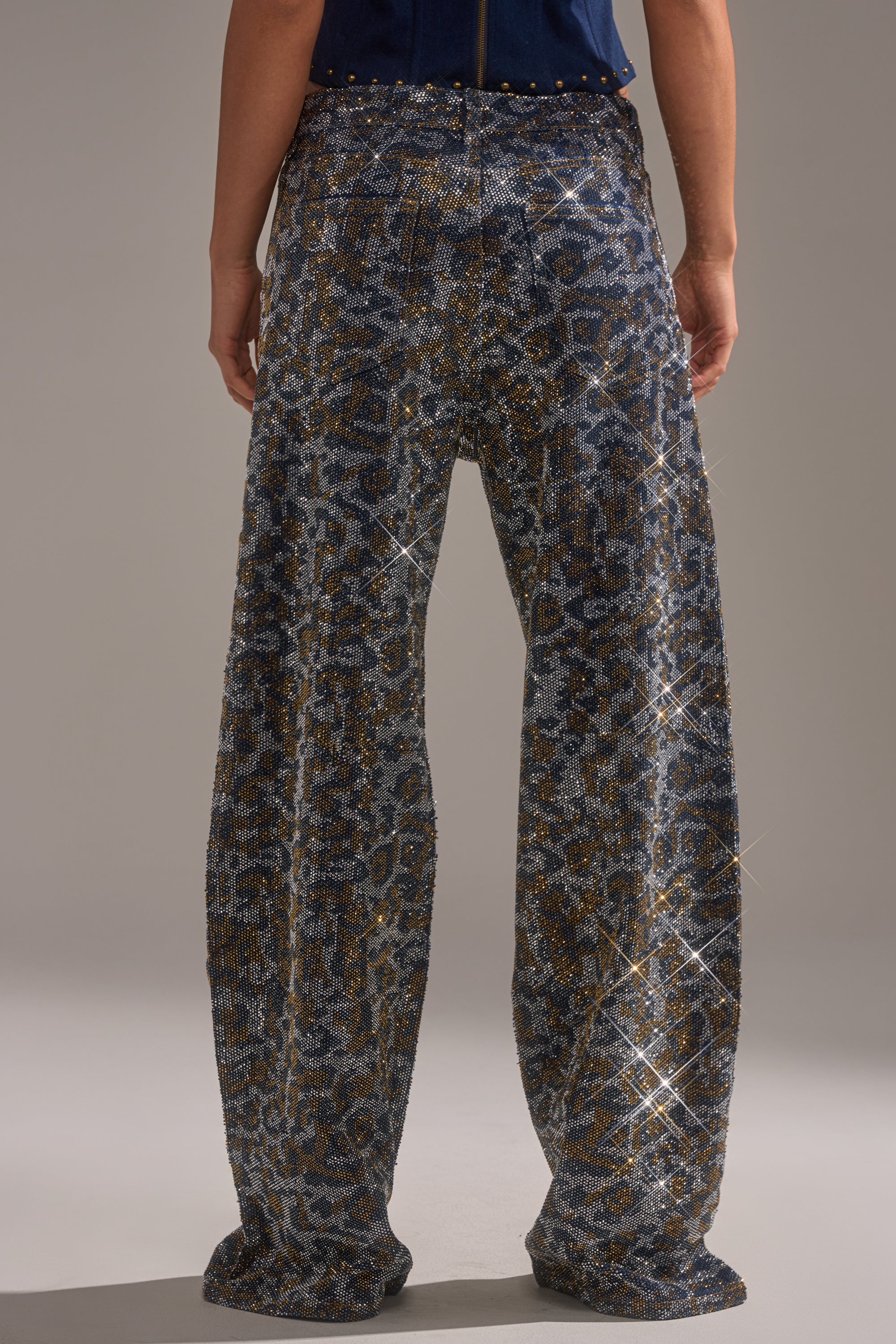 A person stands facing away, wearing the CHEETAH GIRLY RHINESTONE DENIM PANT—loose, shimmering blue and gold wide-leg jeans that reach the floor and sparkle under the light with a glittering effect. 3