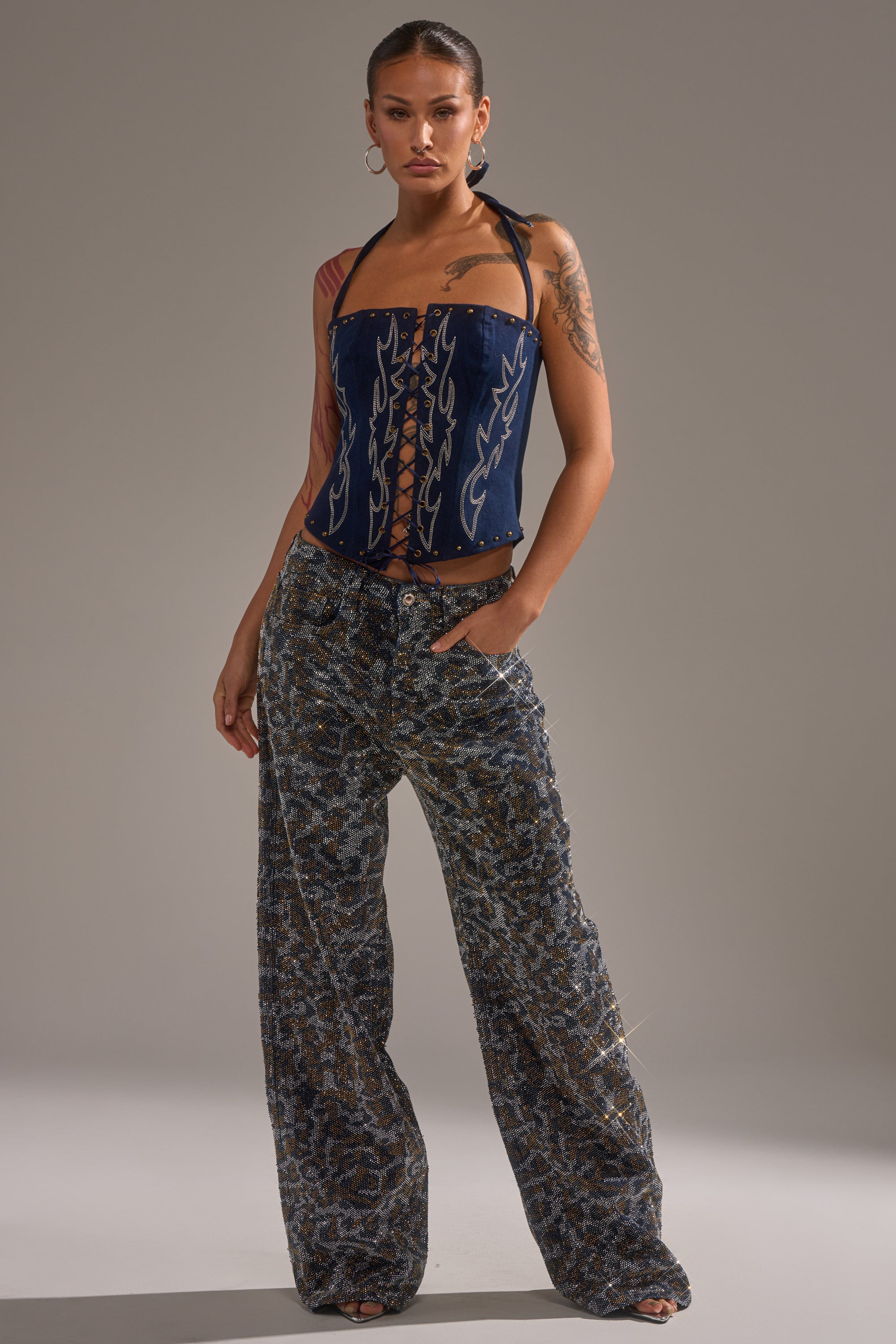 A woman poses confidently in a navy halter corset top and CHEETAH GIRLY RHINESTONE DENIM PANT. She accessorizes with hoop earrings, shows off her tattoos, and has her hair pulled back against a plain gray backdrop. 4