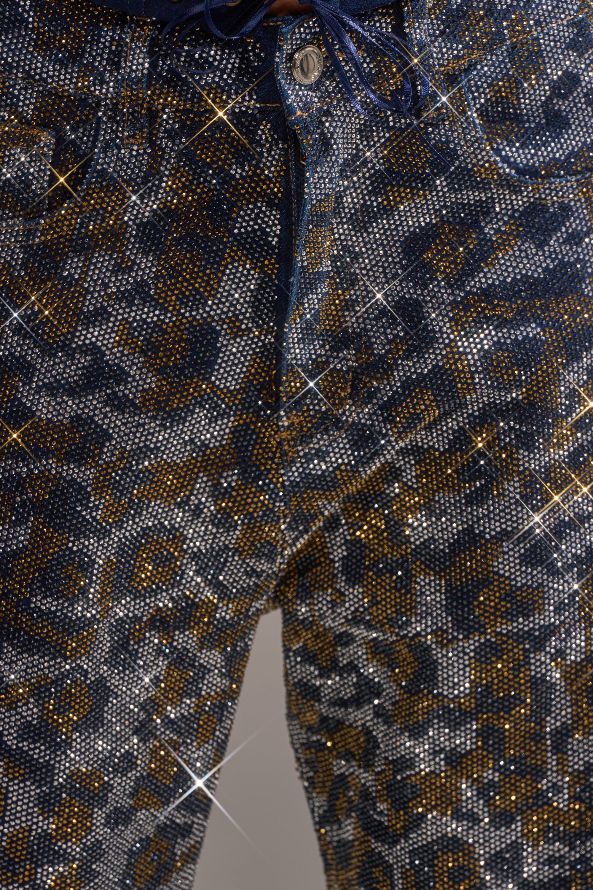 Close-up of the CHEETAH GIRLY RHINESTONE DENIM PANT in blue and gold, high-rise and embellished with sparkling rhinestones that catch the light, creating star-like reflections across the denim. 5