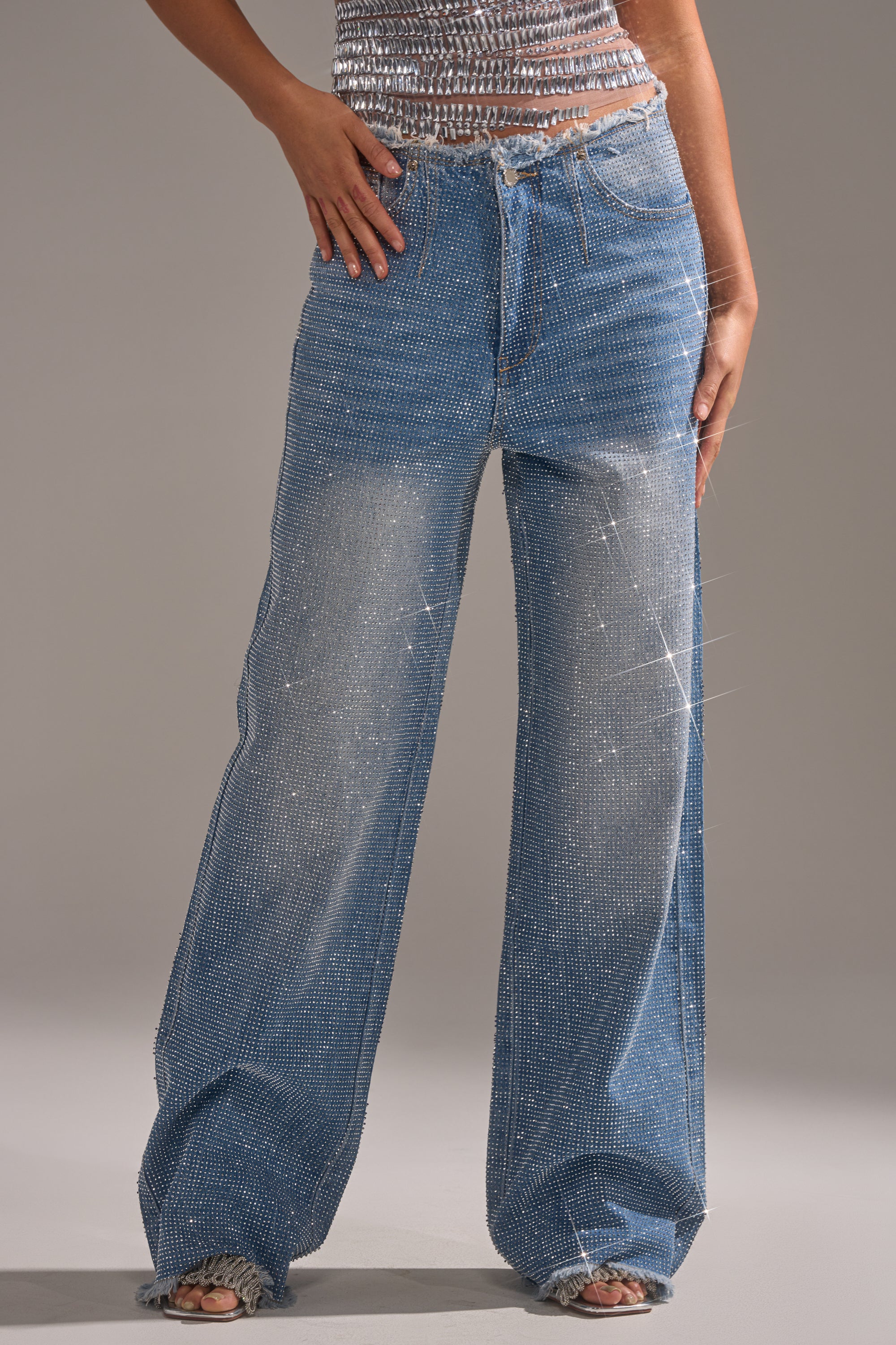 A person wears the FULL MOMENT RELAXED DENIM PANT, featuring a high-waisted, wide-leg fit with rhinestone embellishments, paired with a shiny sleeveless top and stands against a plain gray background. 0