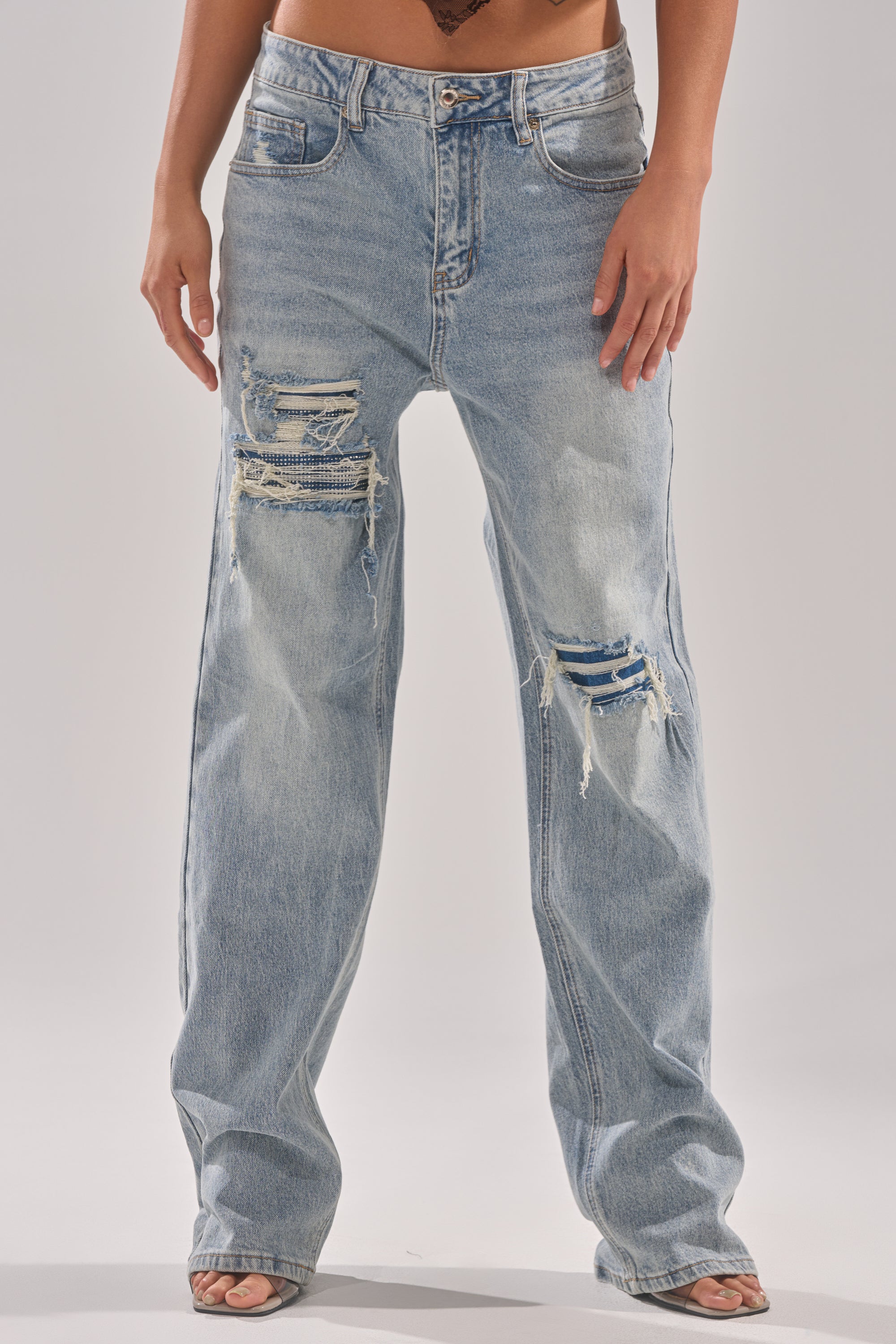 A person wears the RUN AWAY WITH ME RELAXED DENIM—light blue, high-rise jeans featuring multiple distressed and ripped details on the thighs and knees—paired with open-toe shoes for a casual, relaxed look. 1