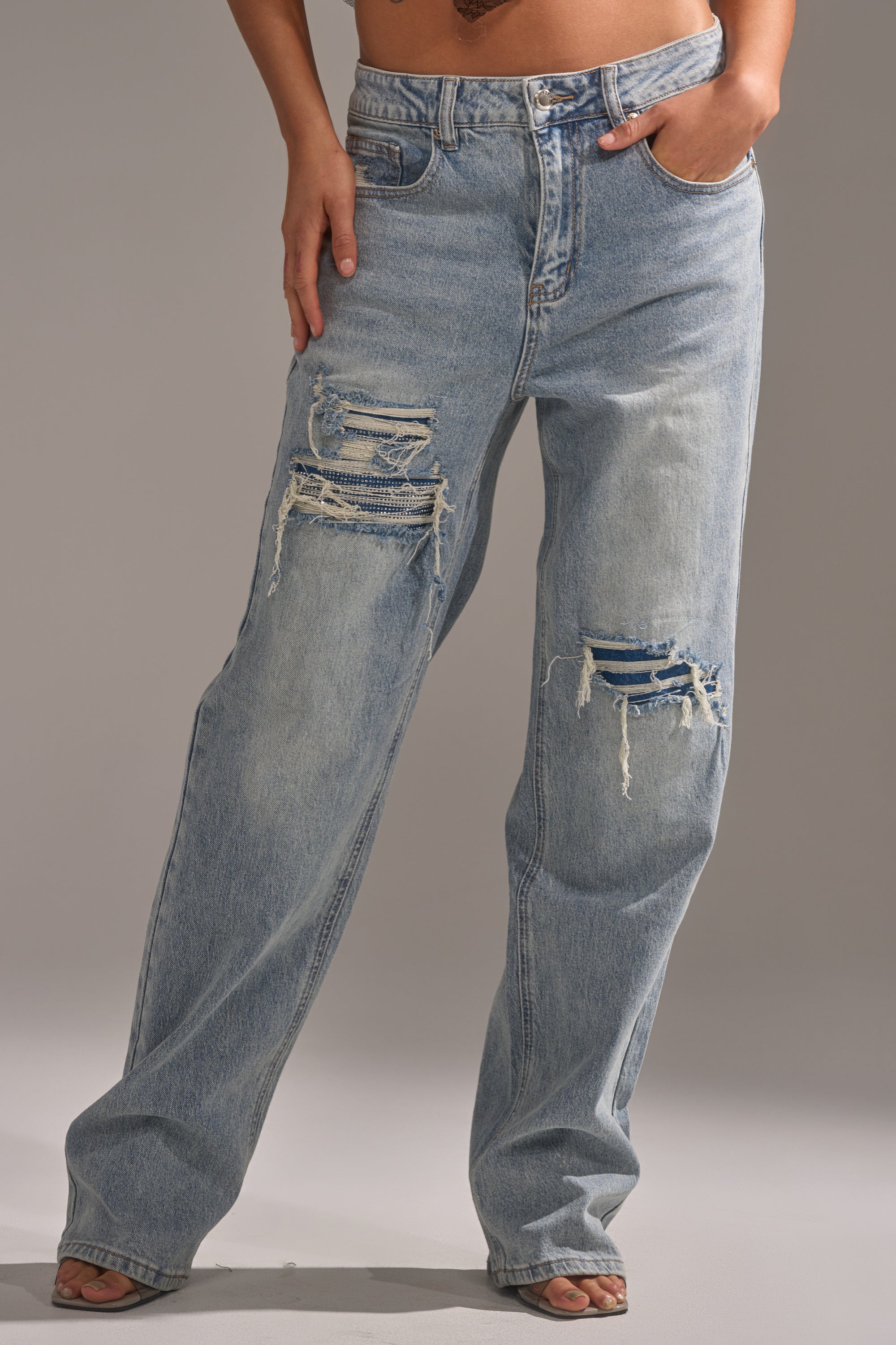 A person models the RUN AWAY WITH ME RELAXED DENIM: light blue, high-rise jeans with a relaxed fit and large ripped, distressed patches on both thighs, set against a plain backdrop. 2