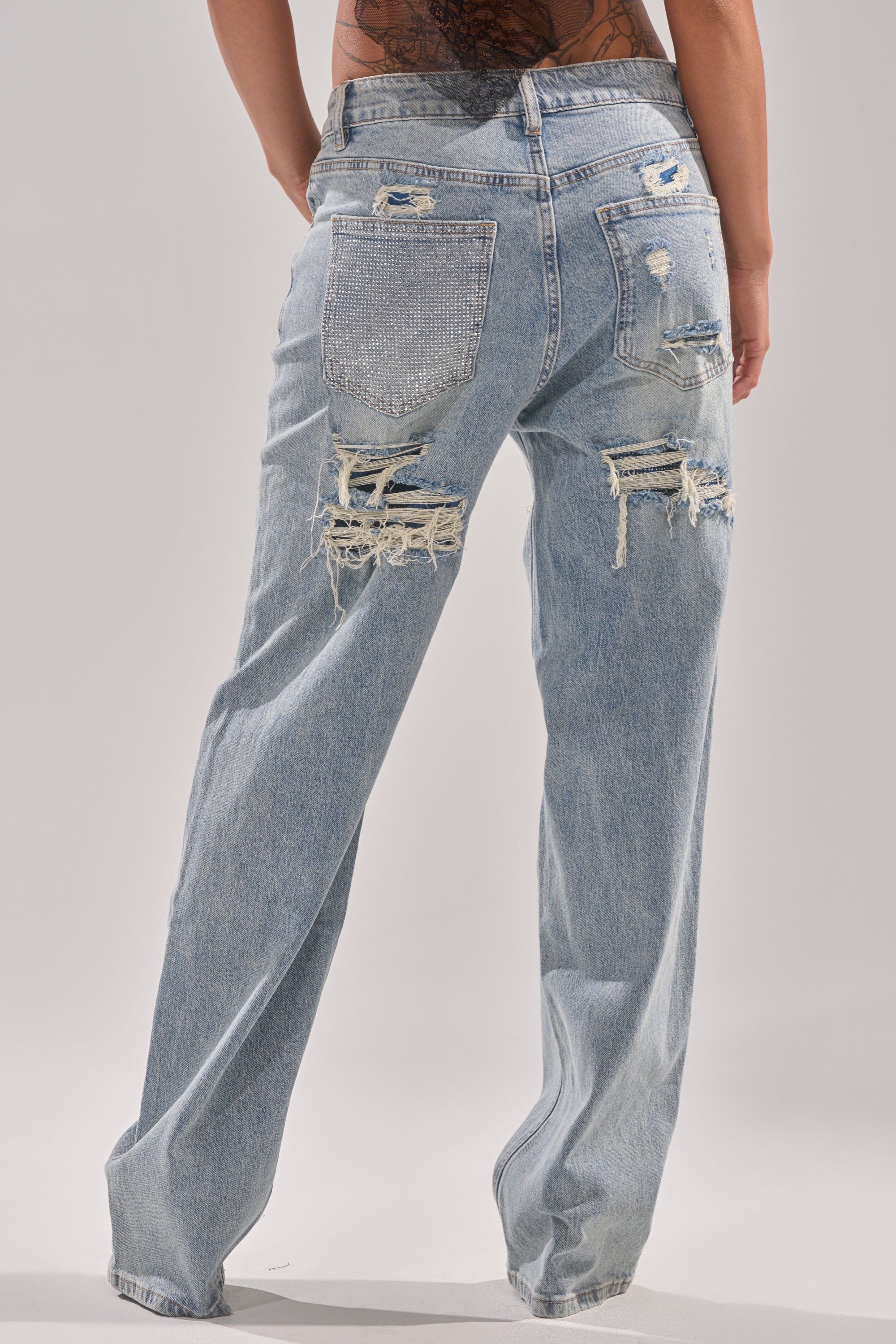 A person shown from the back wears RUN AWAY WITH ME RELAXED DENIM high-rise jeans in light blue, featuring heavy distressing, frayed holes, and a rhinestone embellished back pocket. 4