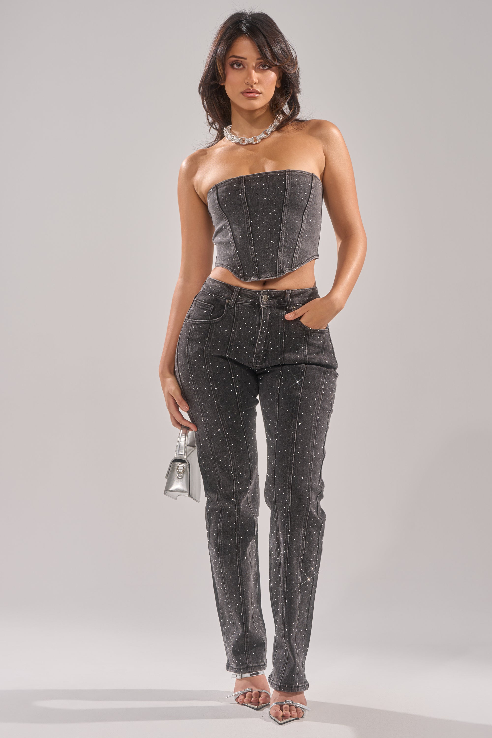 A woman faces forward, wearing a strapless gray top and LETS GET IT RELAXED DENIM PANT with sparkling rhinestone details, silver heels, a chunky necklace, and holding a small silver handbag. 1