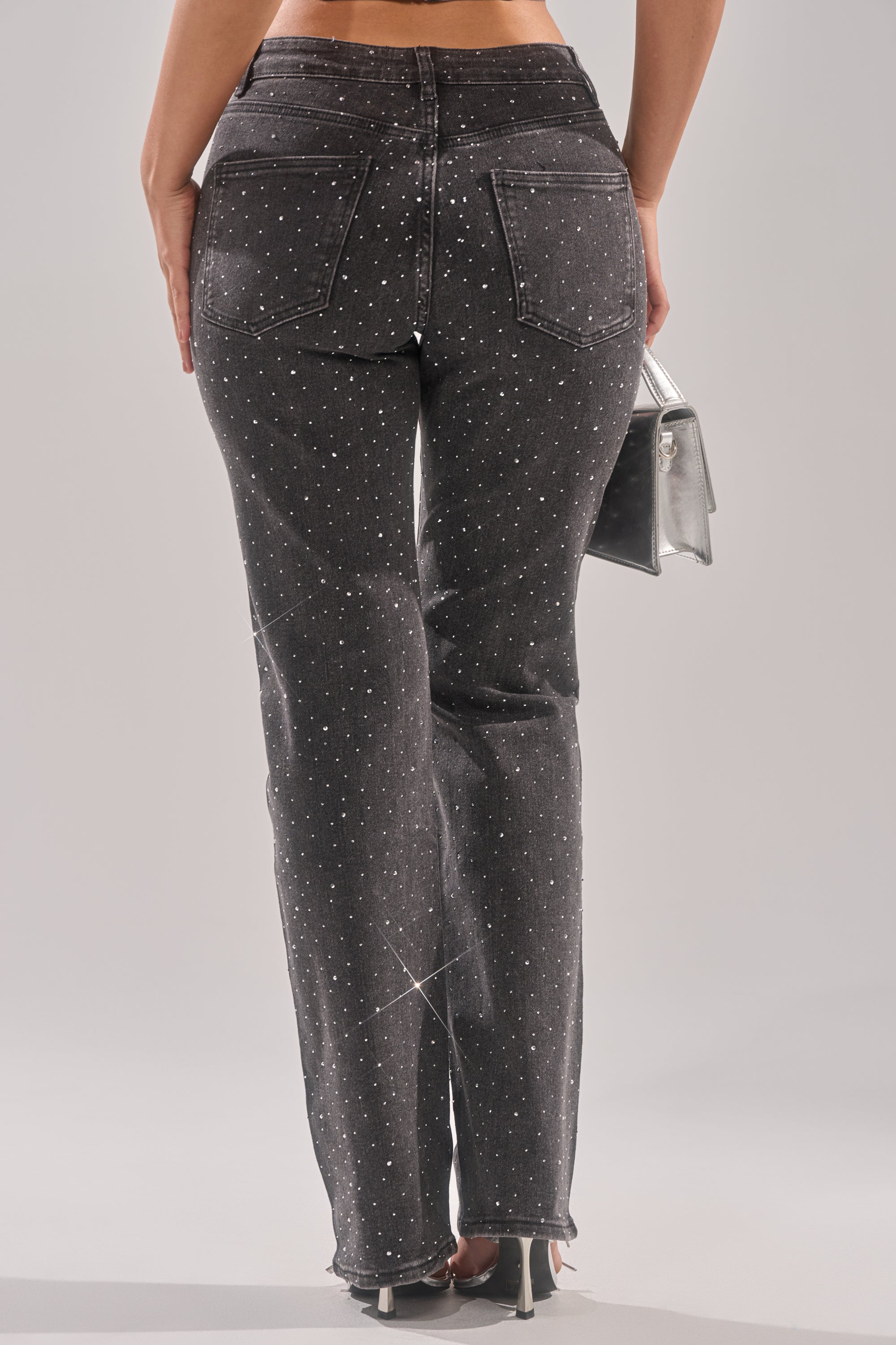 A woman faces away wearing LETS GET IT RELAXED DENIM PANT, high rise black stretch jeans with small sparkling rhinestones, back pockets, a slight flare at the hem, and carrying a silver handbag. 5