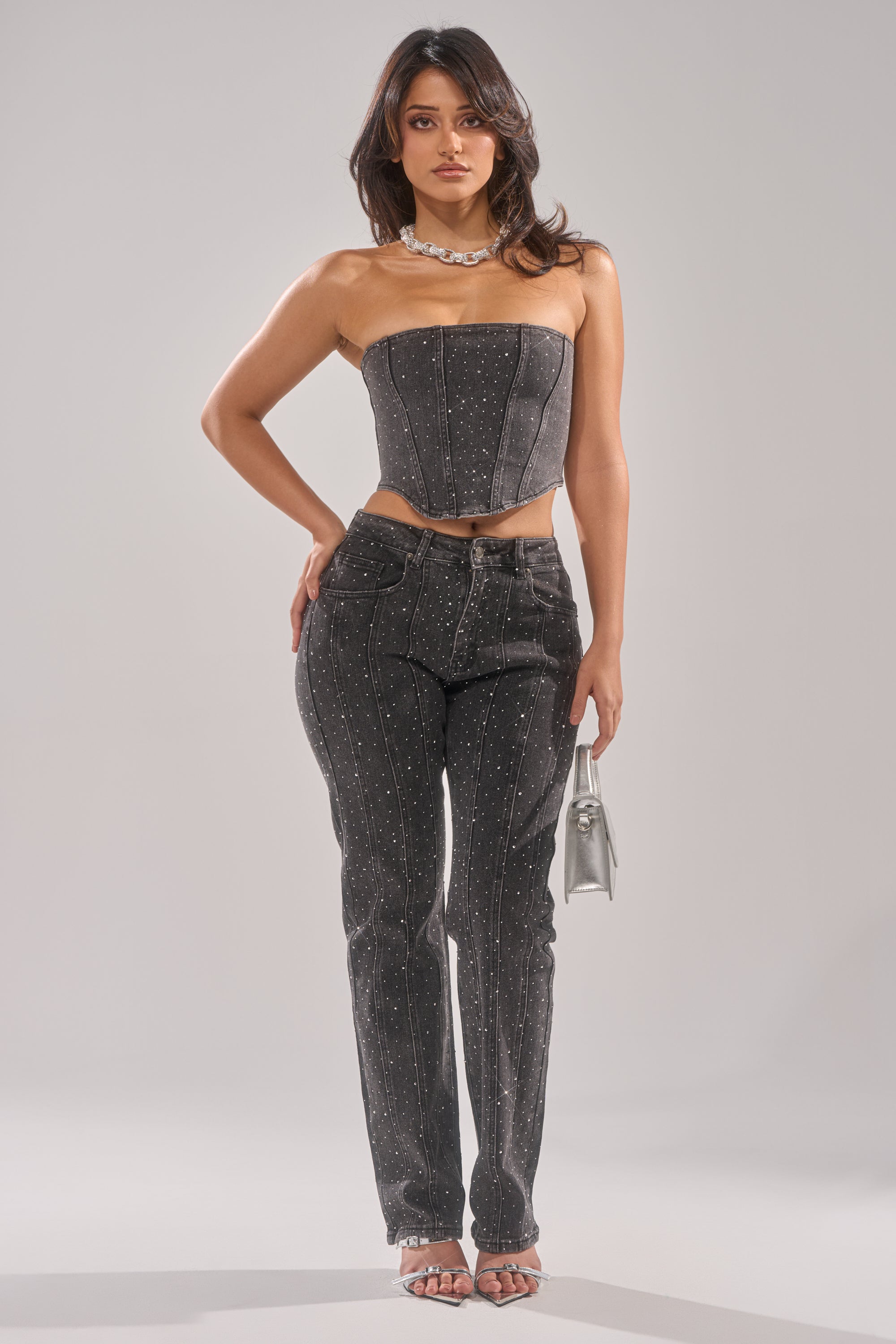A woman poses in the LETS GET IT DENIM CORSET TOP, a strapless, rhinestone-embellished, fitted denim piece styled with matching high-waisted pants, heels, a necklace, and a silver handbag against a plain background. 5