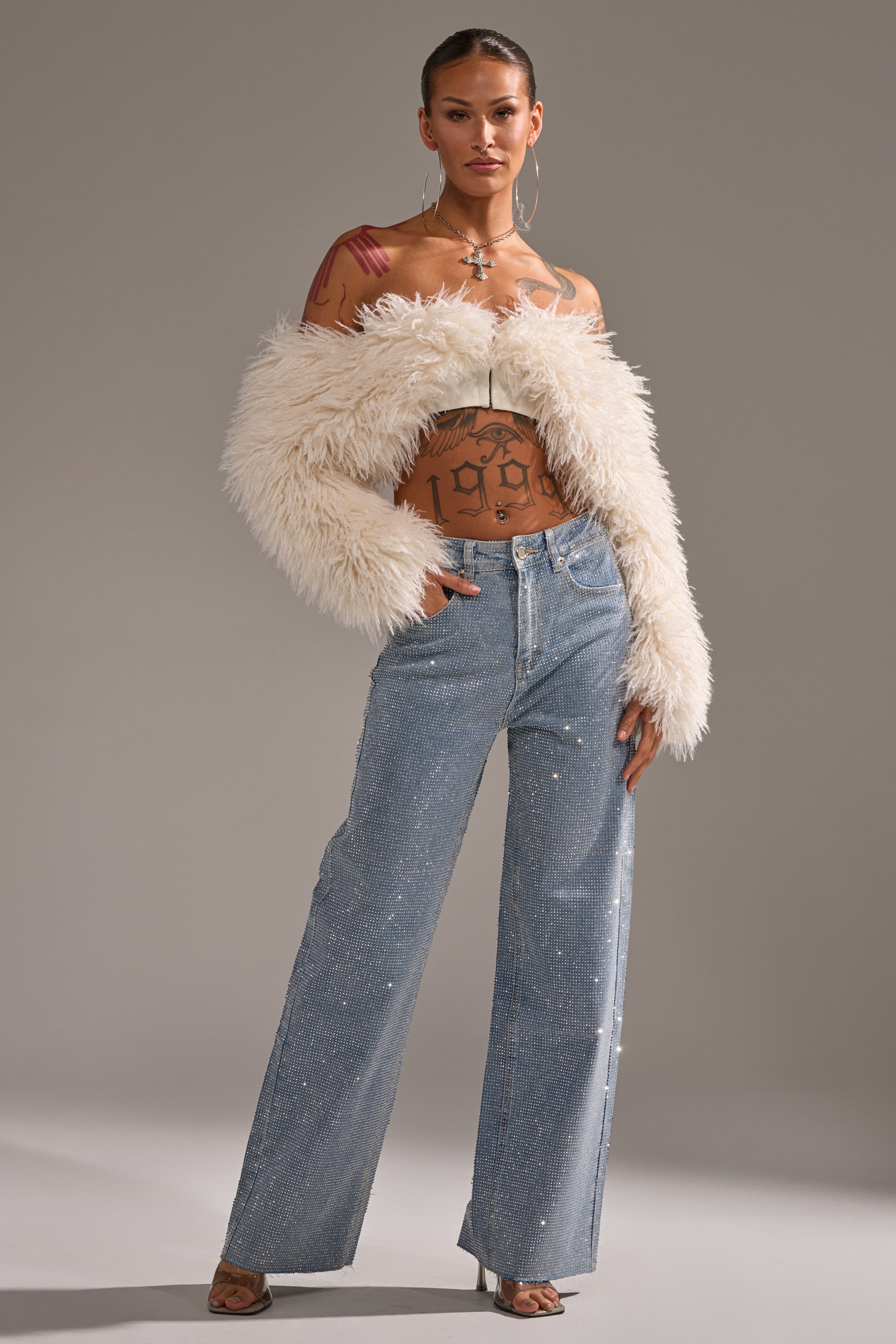 A woman poses in DANCING QUEEN RELAXED DENIM high-waisted wide-leg jeans, a cream faux fur shrug, and clear heels. She accessorizes with hoop earrings, layered necklaces, and reveals tattoos on her torso and neck against a gray backdrop.