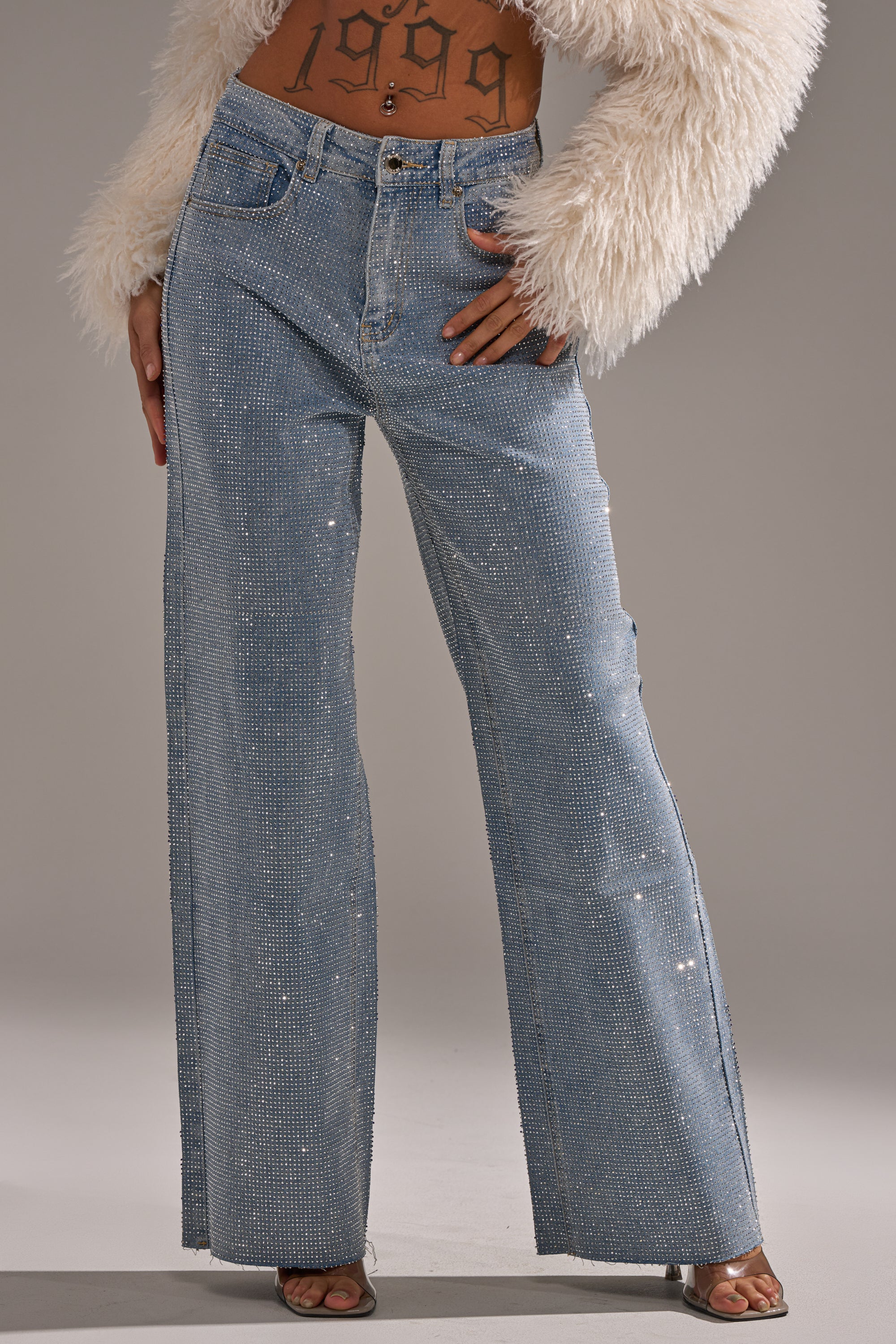 A person in DANCING QUEEN RELAXED DENIM—sparkling, rhinestone-embellished wide-leg jeans—paired with a furry white jacket and open-toed shoes, "1999" tattooed on their midriff. Background: plain gray.
