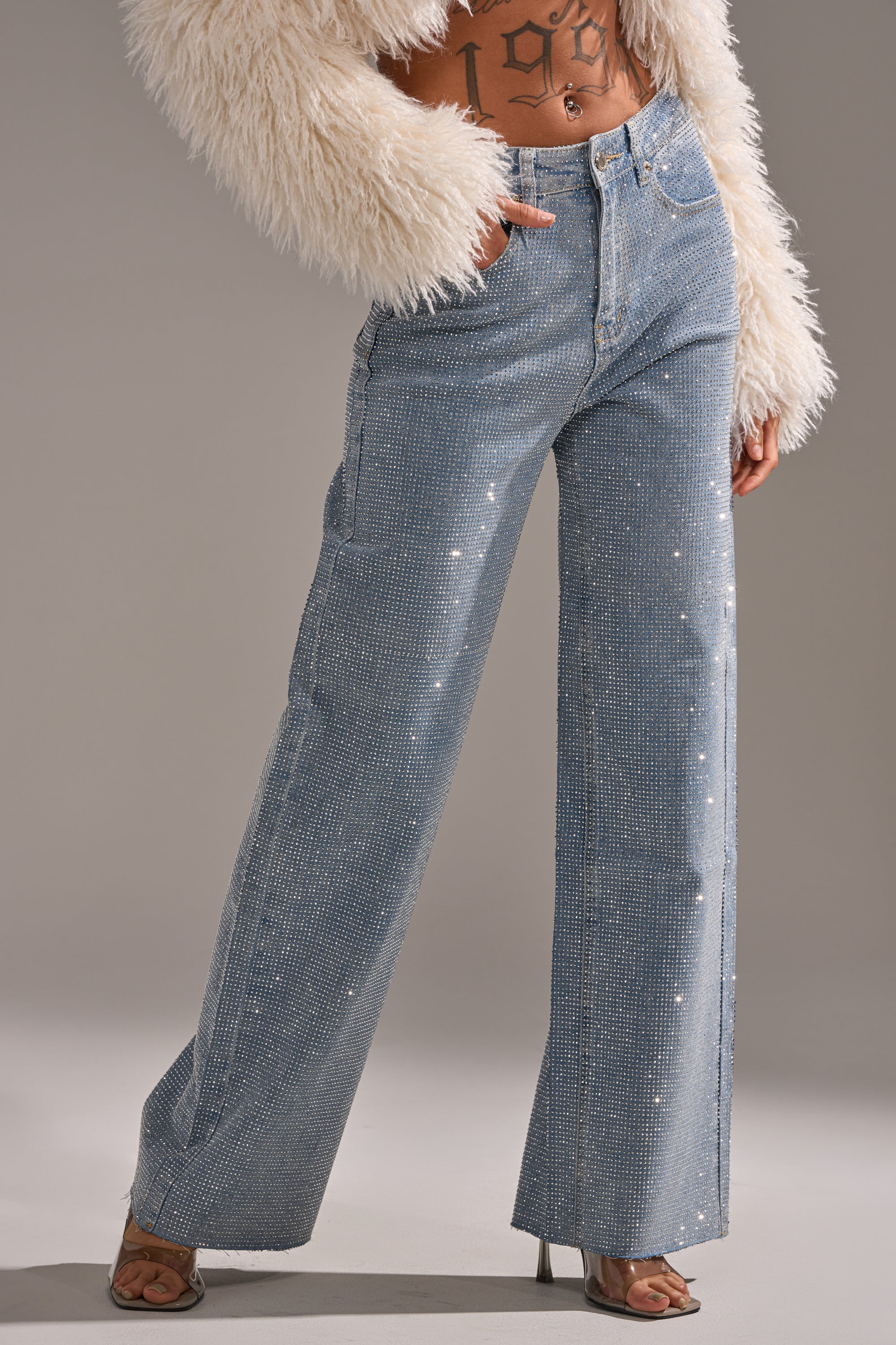 Wearing DANCING QUEEN RELAXED DENIM jeans with rhinestone embellishments, a white faux fur jacket, and open-toe heels. Their tattooed midriff adds edge against the plain, neutral background.