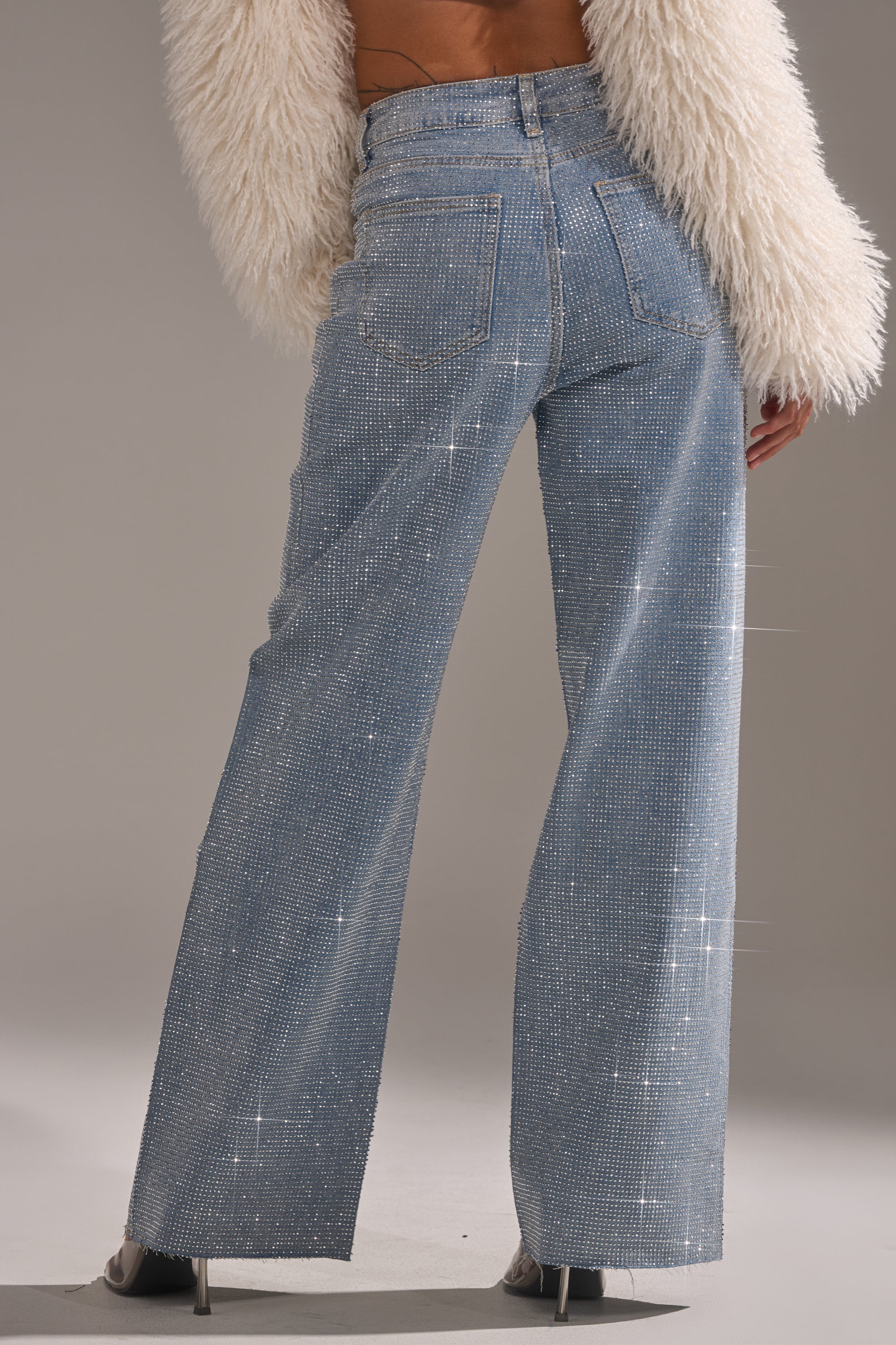 Wearing the DANCING QUEEN RELAXED DENIM, a person stands with their back to us, showcasing wide-leg blue jeans adorned with rhinestones and paired with a white fluffy jacket that sparkles under the light.