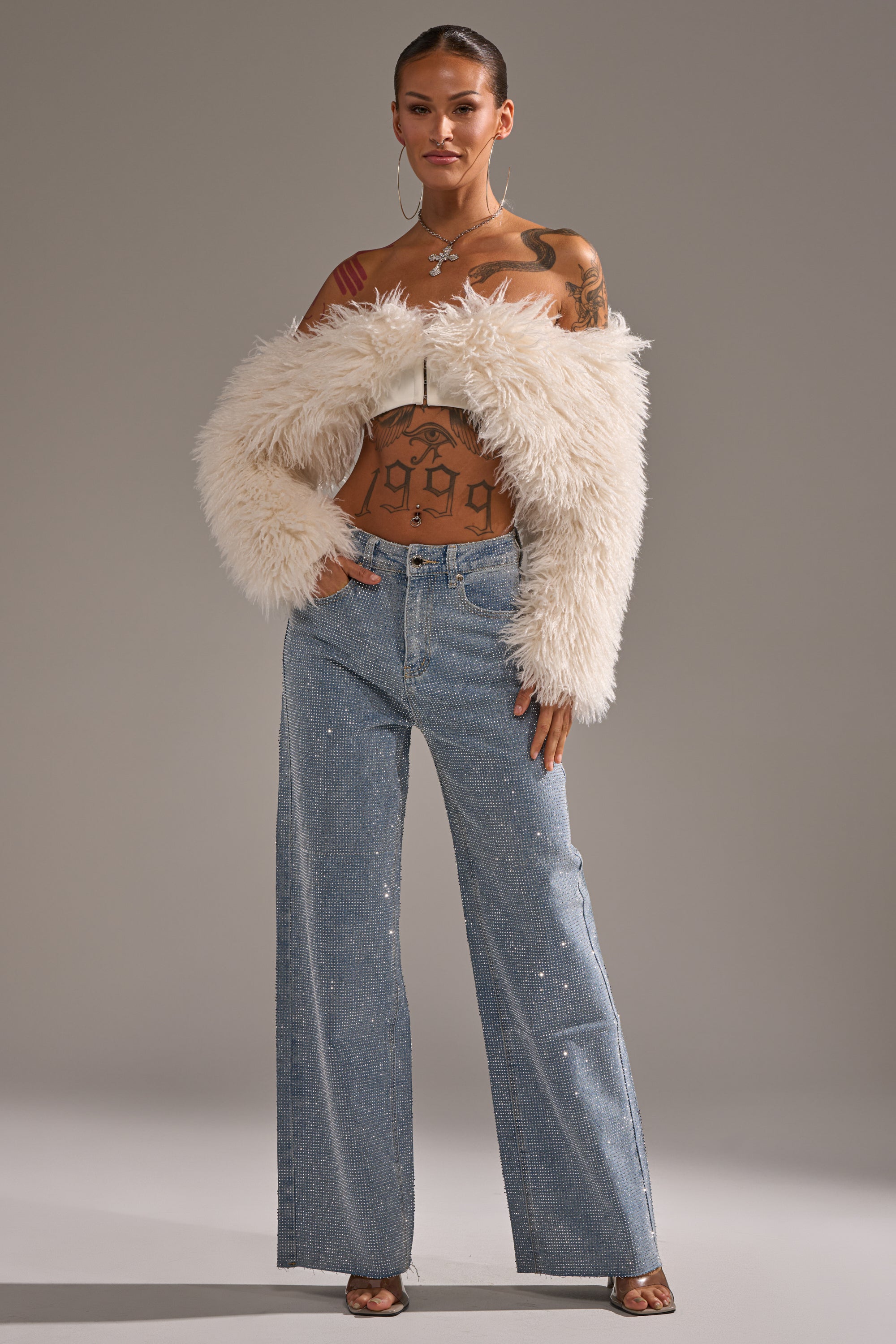 Wearing DANCING QUEEN RELAXED DENIM with a wide-leg fit, this person styles the light blue jeans with a white furry off-the-shoulder jacket and open-toed heels, hair pulled back to reveal tattoos on their chest, arms, and torso.