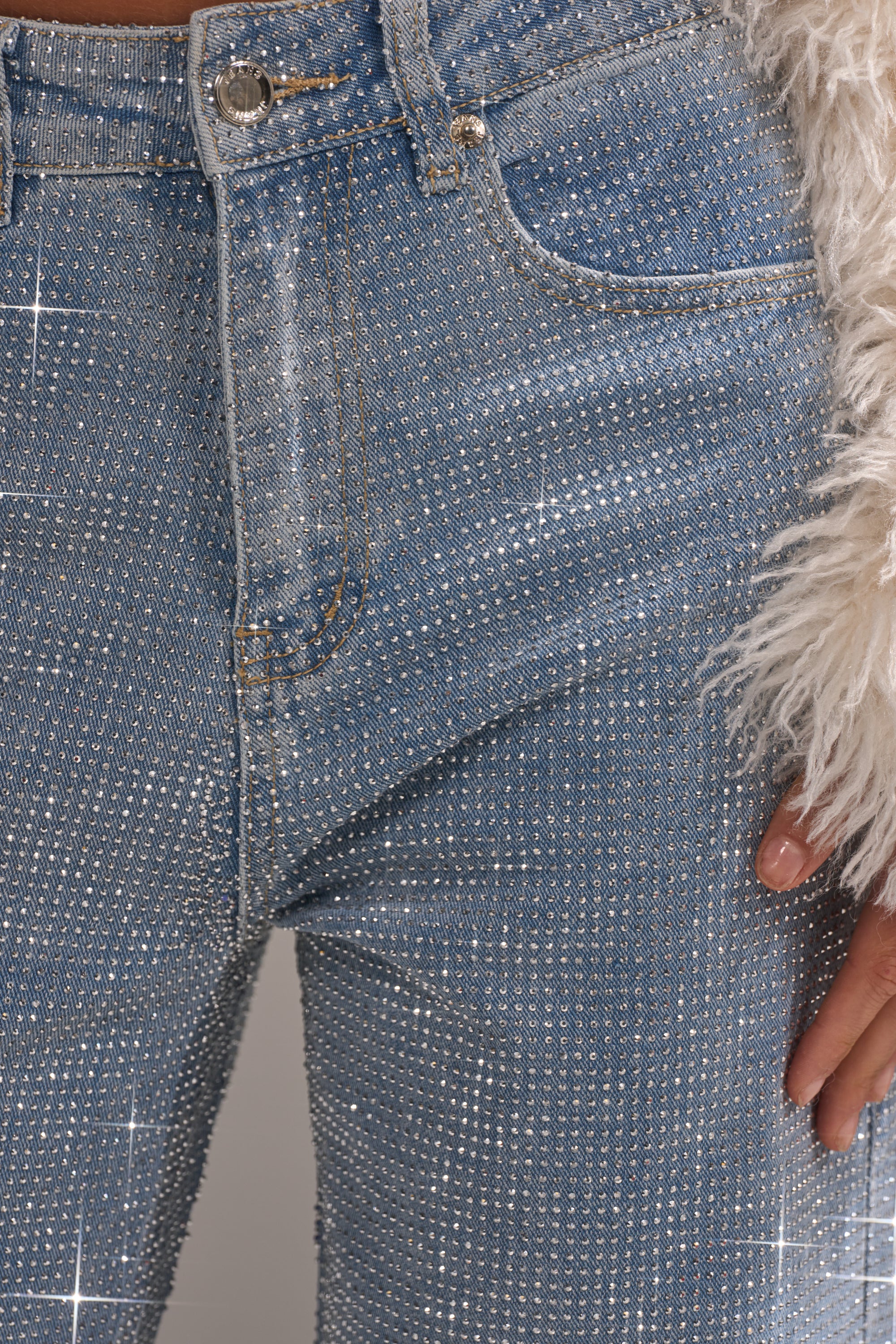 Close-up of a hand near the pocket of DANCING QUEEN RELAXED DENIM—sparkly blue jeans with rhinestone embellishments and a fuzzy white jacket. The image highlights the jeans’ textured shimmer and relaxed feel.