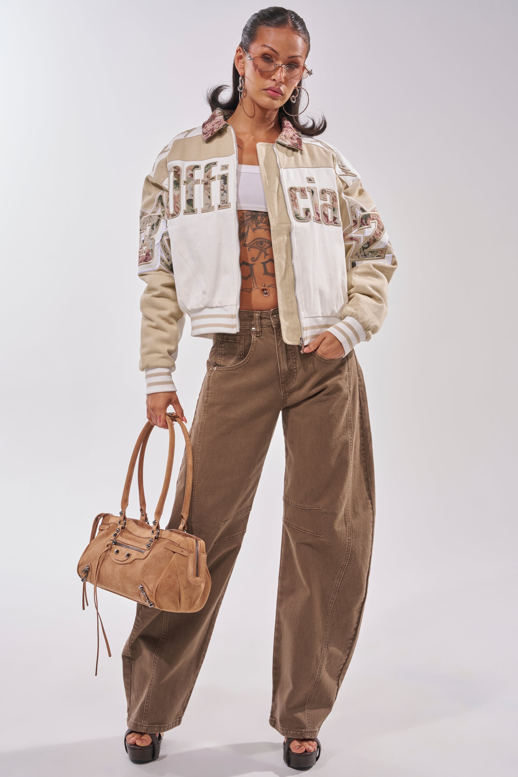 A woman with sunglasses, hoop earrings, and visible midriff tattoos wears FEELING GREAT BARREL DENIM jeans, a white and beige varsity jacket, white crop top, black heels, and holds a tan handbag as she poses confidently. 0