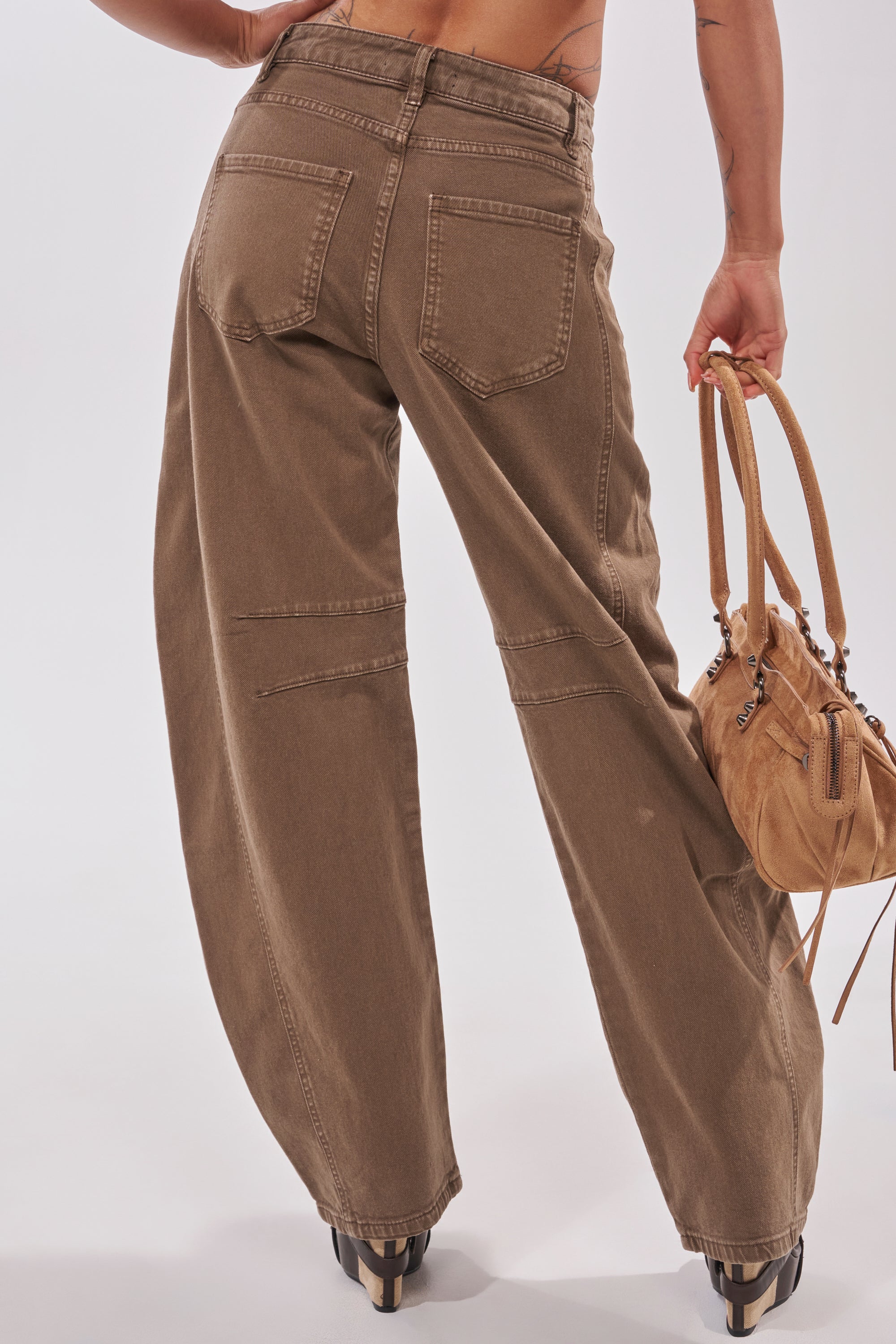 A person is shown from the back wearing FEELING GREAT BARREL DENIM—loose-fitting brown jeans with a barrel leg silhouette, large back pockets, and visible seam details—holding a tan handbag in their right hand. 4