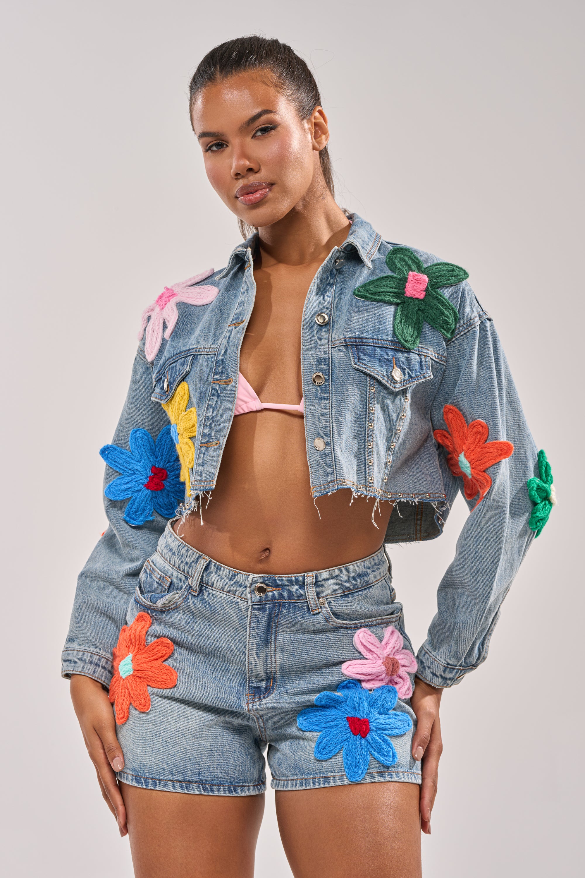 A woman models the FLOWER PATCH DENIM JACKET with floral appliques, pairing it with distressed-hem shorts and a light pink bikini top, striking a confident pose against a plain light background. 1