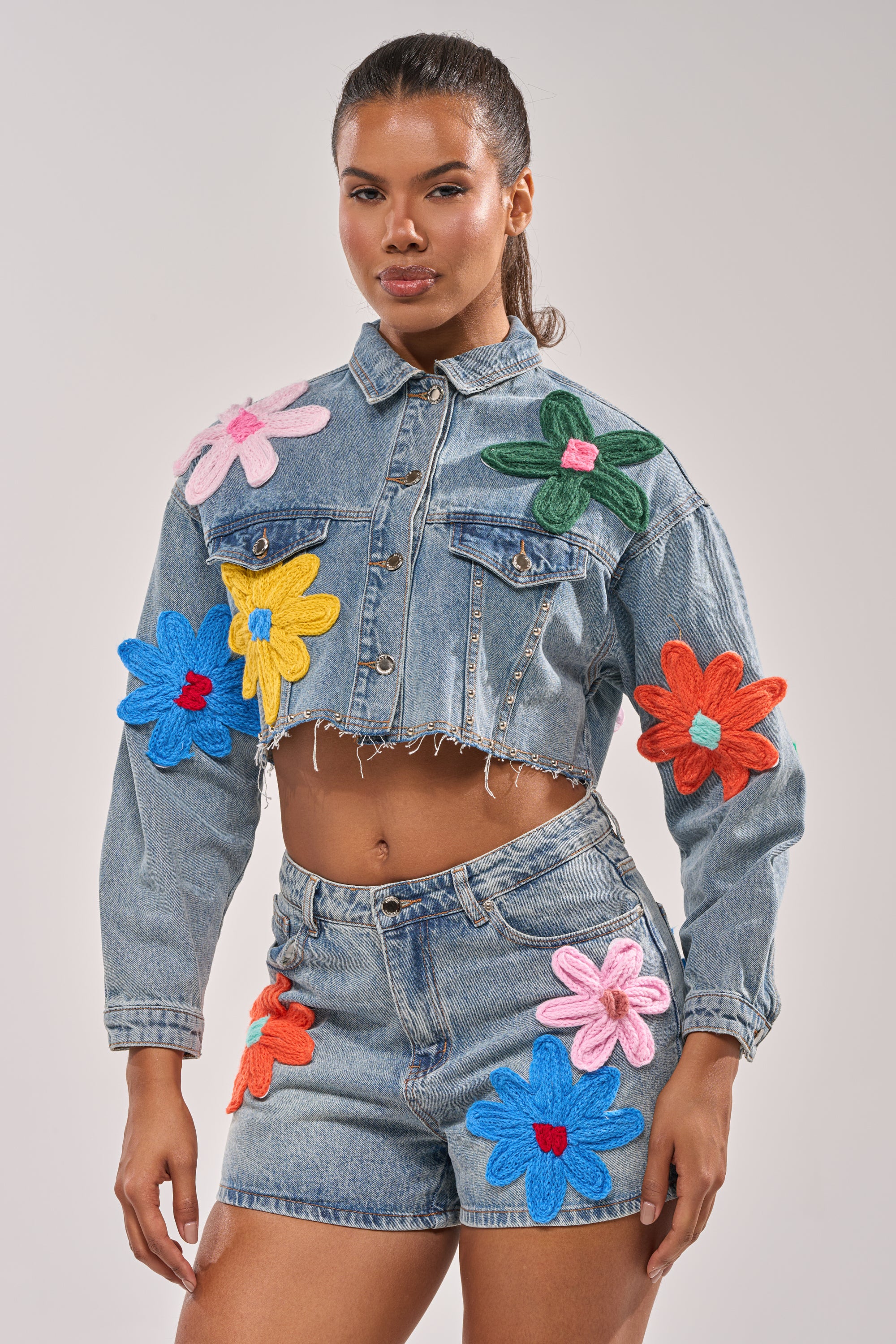 A woman confidently models the FLOWER PATCH DENIM JACKET, featuring a cropped fit and distressed hem, paired with high-waisted shorts decorated with colorful floral appliques against a plain background. 2