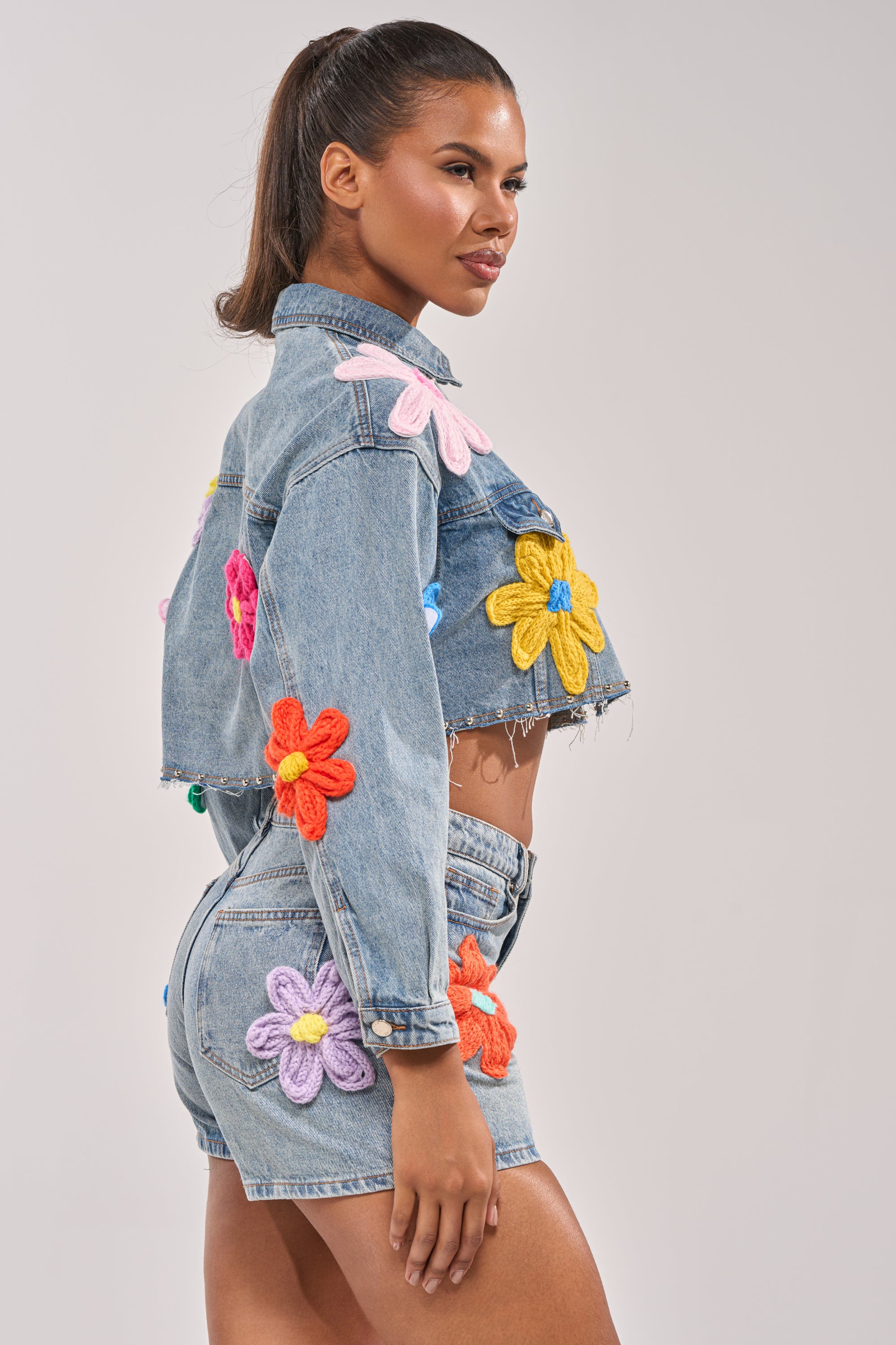 A woman models the FLOWER PATCH DENIM JACKET, a cropped denim piece with a distressed hem and bold, colorful floral appliqués, paired with matching shorts as she stands confidently against a plain light background. 4