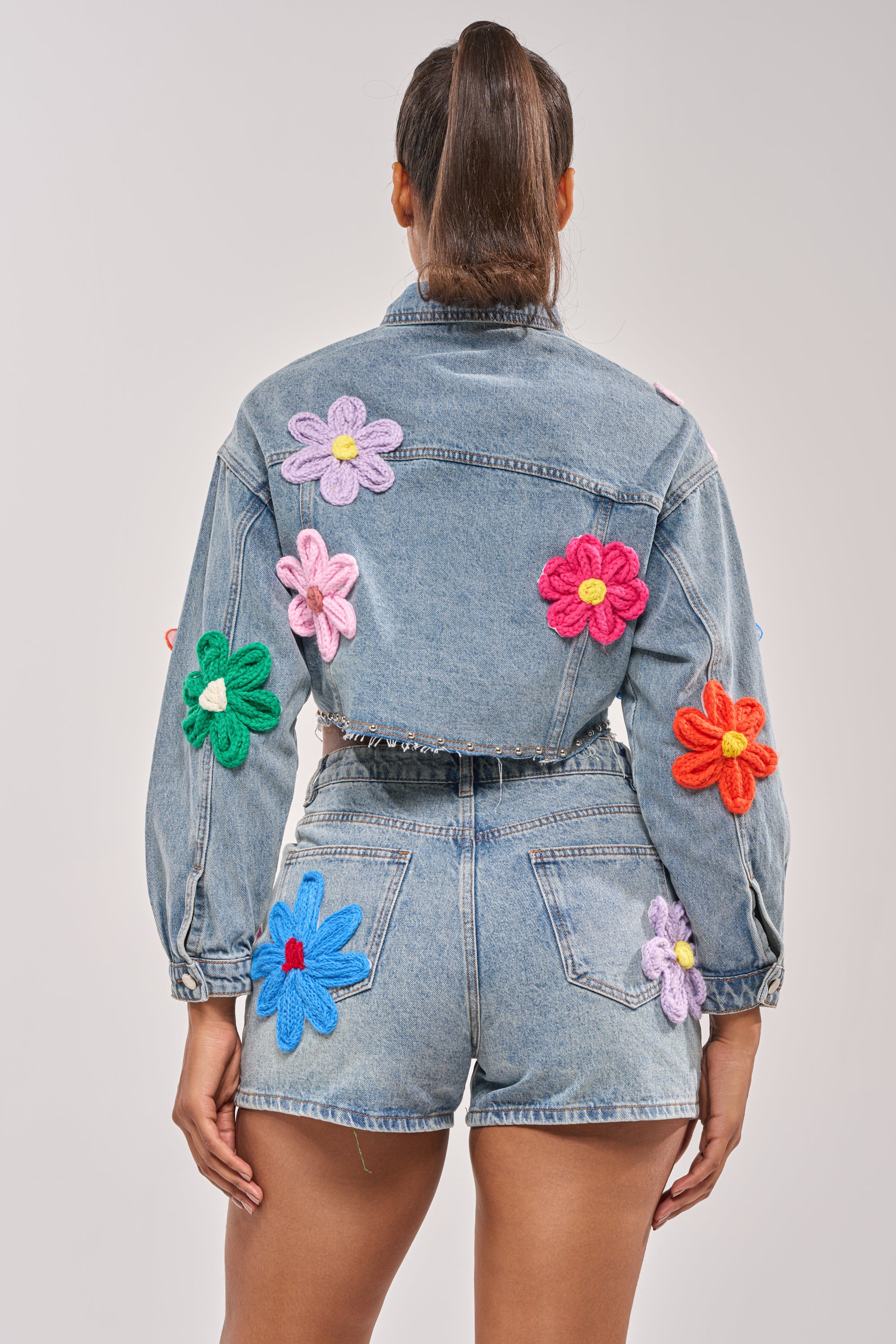 A person with a high ponytail is seen from the back wearing the FLOWER PATCH DENIM JACKET with large floral appliques and a cropped distressed hem, paired with matching shorts, against a plain light background. 5