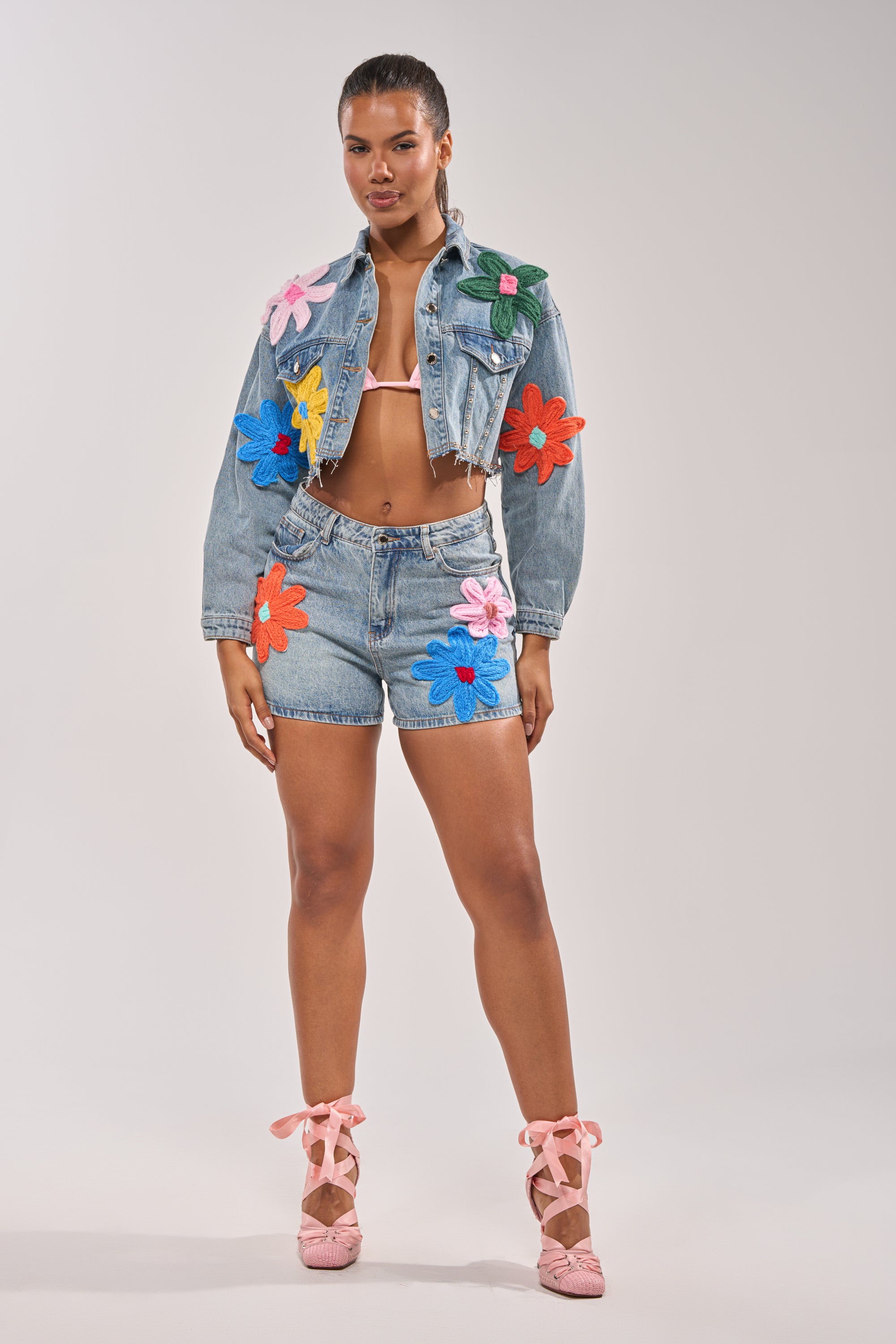 A woman stands confidently in the FLOWER PATCH DENIM JACKET with a cropped distressed hem, paired with matching floral-appliqued shorts, a white bra top, and pink ballet flats against a plain light background. 6