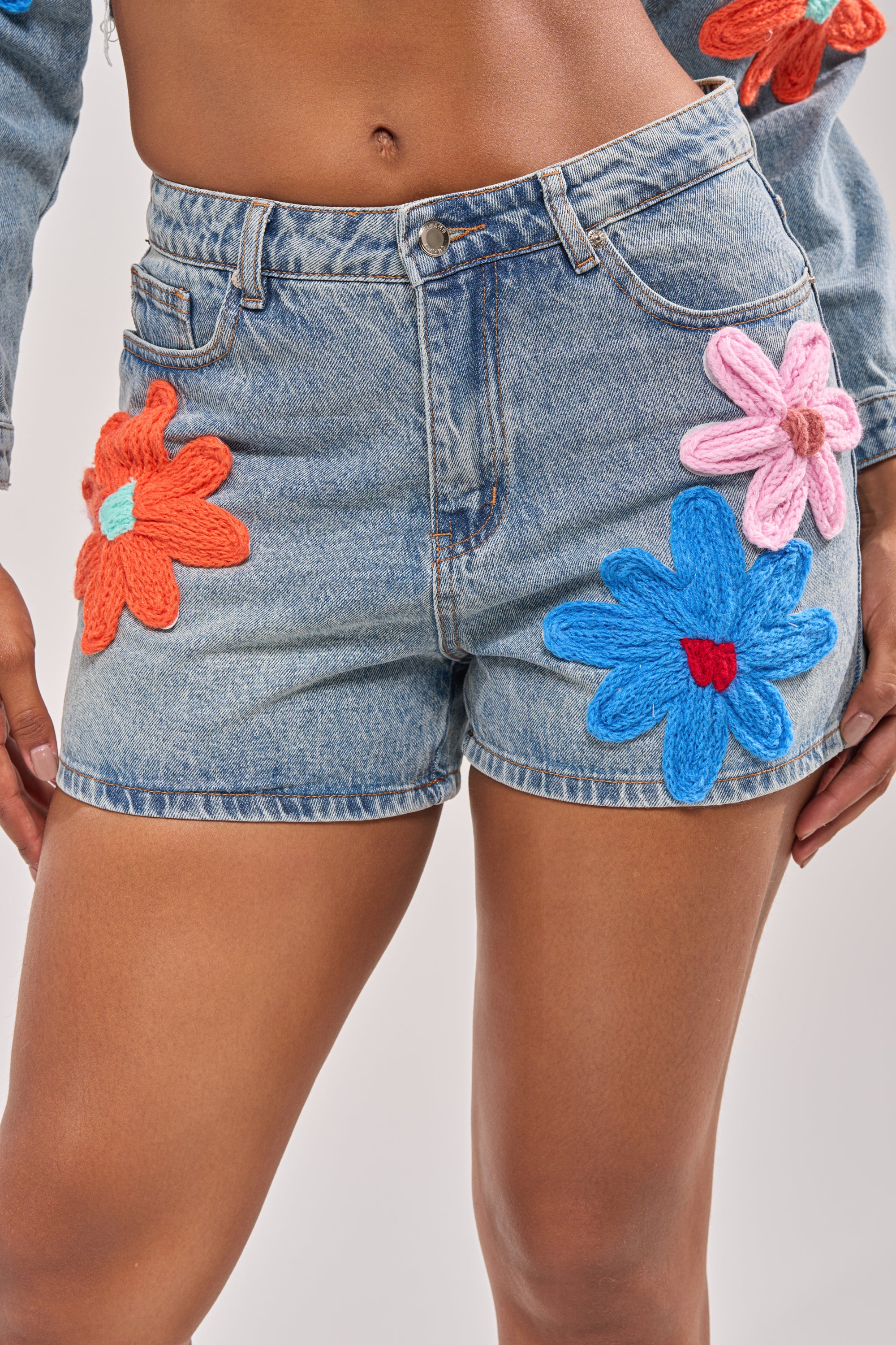 A person wears the FLOWER PATCH DENIM SHORT, a high-rise blue denim short featuring large orange, blue, and pink floral appliques. The photo is cropped above the waist and at mid-thigh. 0