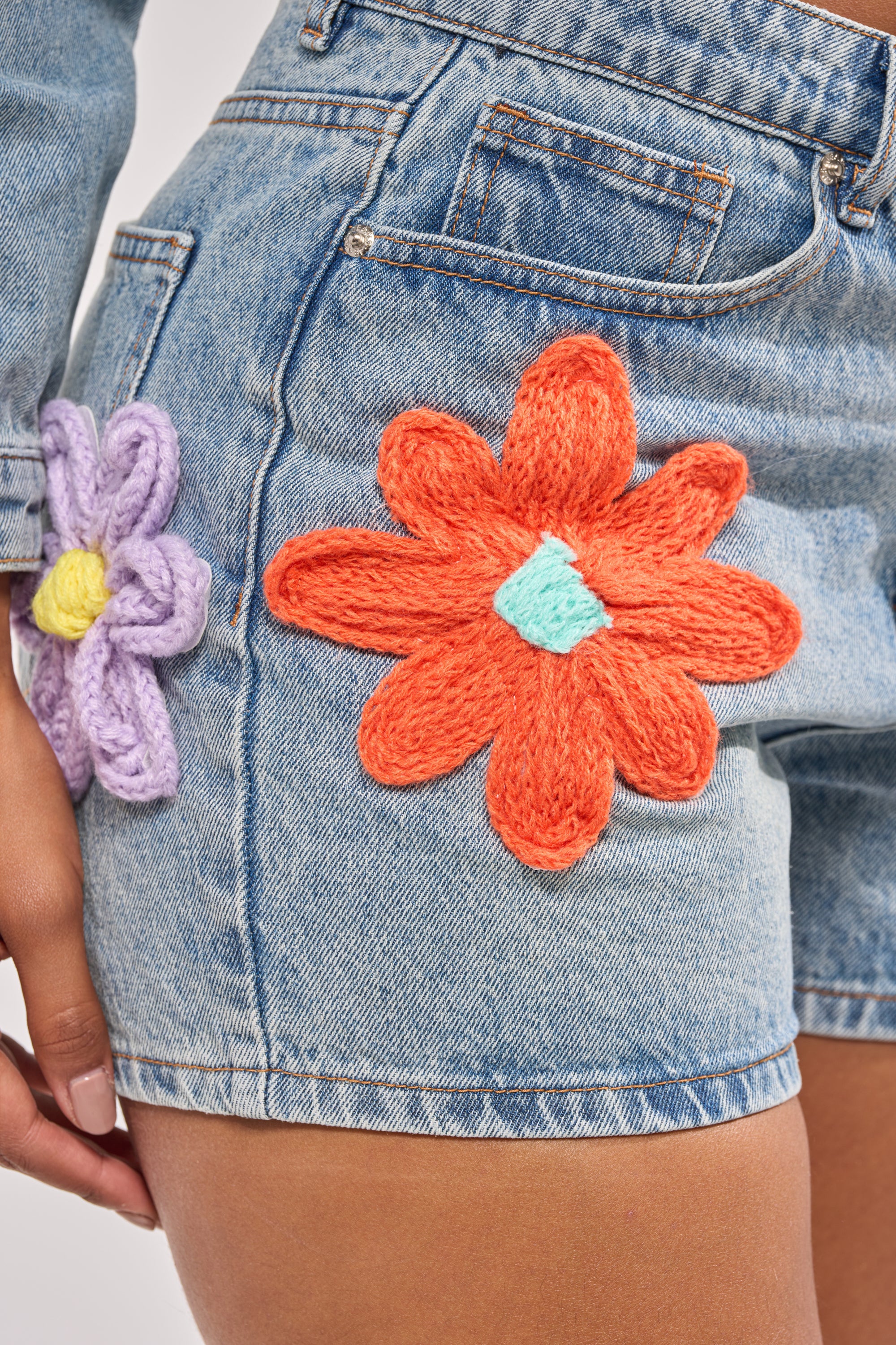 Close-up of a person wearing the FLOWER PATCH DENIM SHORT, featuring large, colorful floral appliques—one orange with a blue center and one purple with a yellow center—on the front and side. The person’s hand rests near the shorts. 8