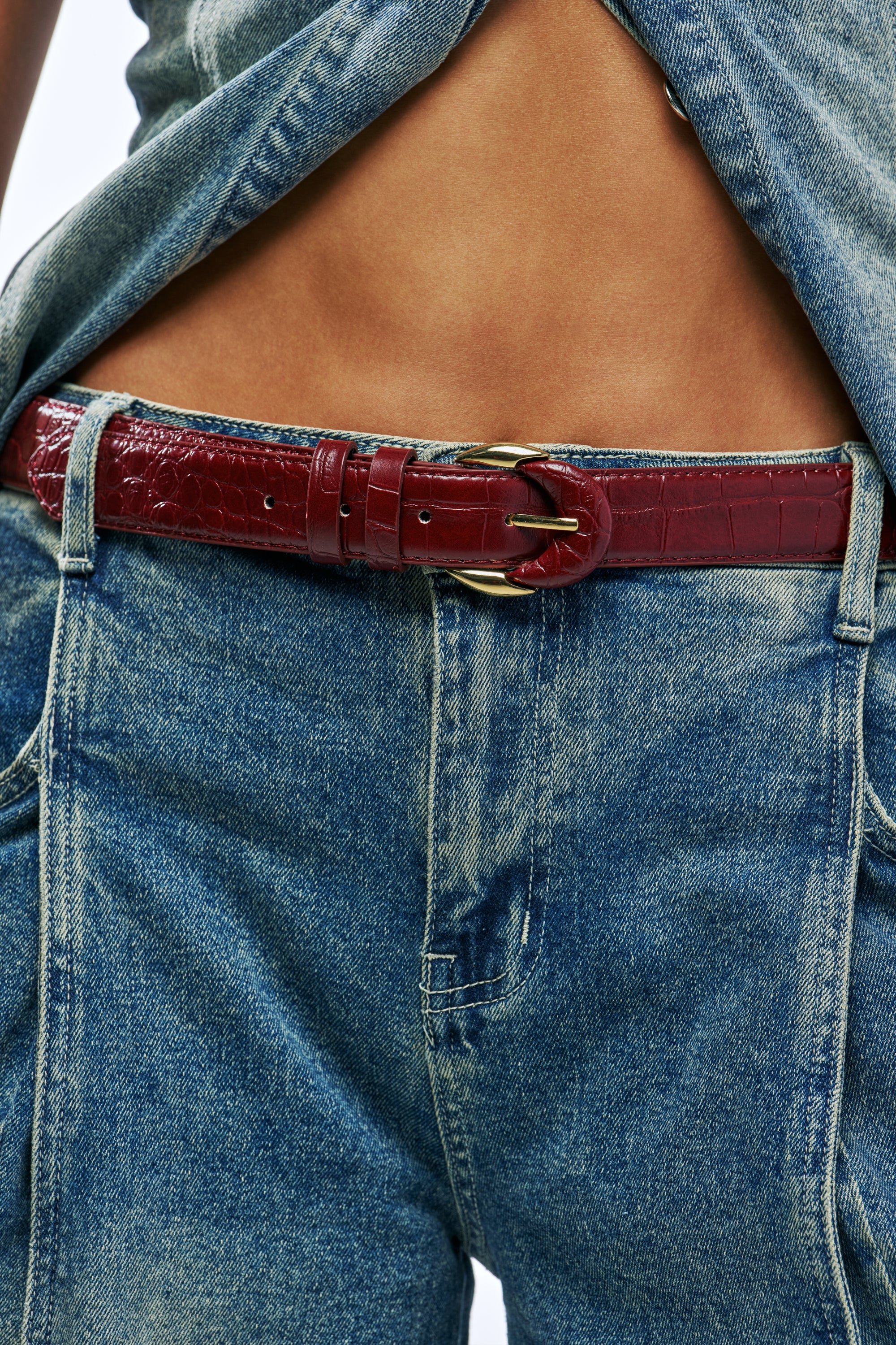 A person wearing a burgundy faux leather CROCODILE QUEEN BELT with a gold buckle over blue jeans.