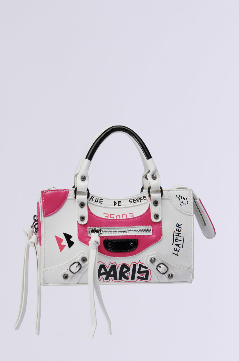 The "PARIS LOVES ME GRAFFITI PURSE" is a white faux leather handbag featuring black and pink details with Paris-inspired graphics in graffiti-style text and symbols. It includes a small top handle, zipper accents, and stylish elements like "Paris" and "Rue de Sevres" written on it.