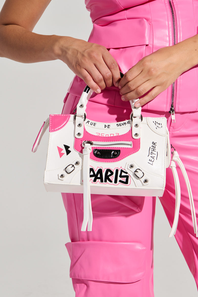 Clad in a pink leather ensemble, an individual holds the PARIS LOVES ME GRAFFITI PURSE, a white faux leather handbag embellished with Paris-themed graphics. The bag showcases black and pink graffiti-style text that reads "PARIS" and "RUE DE SEVRES," enhanced by decorative studs and zippers for an audacious finish.