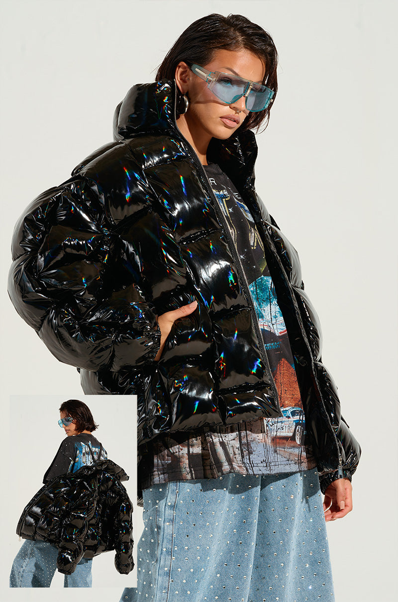 A person confidently poses wearing the BOB AND WEAVE OIL SLICK PUFFER, featuring a metallic front zipper, paired with large blue-tinted sunglasses. They sport loose, faded jeans adorned with sparkly embellishments and have a graphic tee visible under the jacket. Their hair is short and styled back.