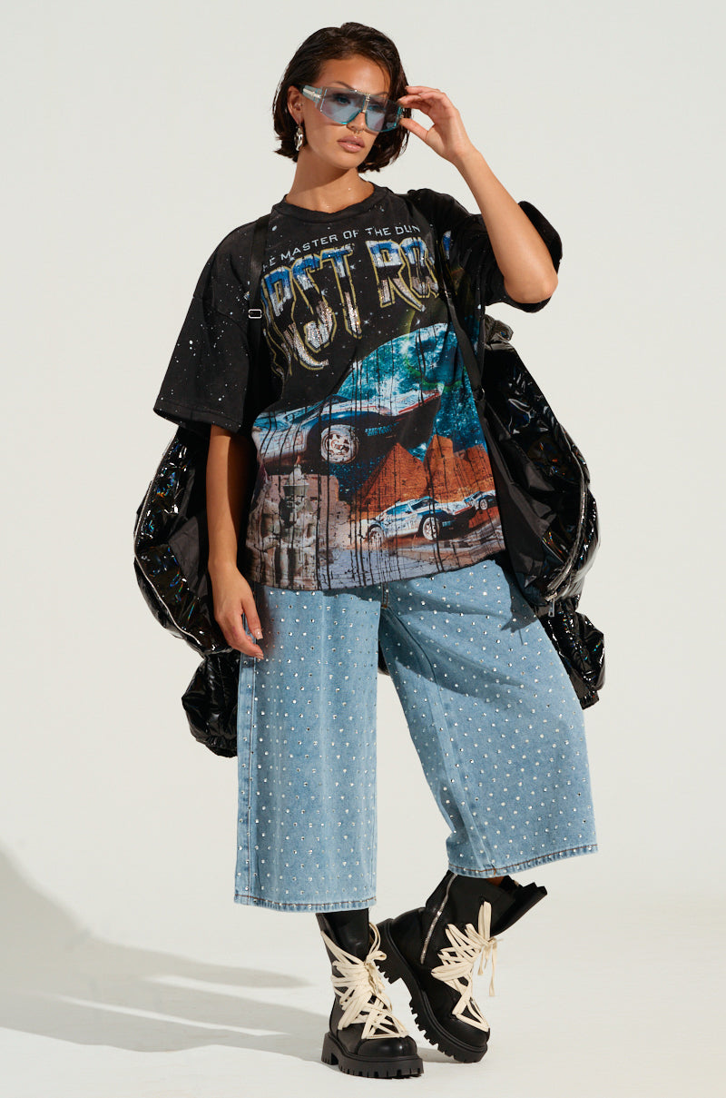 A person styled in oversized sunglasses, a black graphic T-shirt, blue denim culottes, and chunky black boots accented with white laces is wearing the BOB AND WEAVE OIL SLICK PUFFER featuring a metallic front zipper. They strike a pose against a plain background.