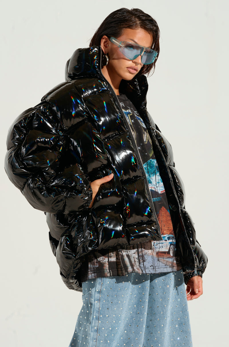 A person wearing the Bob and Weave Oil Slick Puffer, featuring a metallic front zipper, oversized sunglasses, a graphic T-shirt, and wide-leg jeans adorned with small embellishments. They are posing confidently against a neutral background.