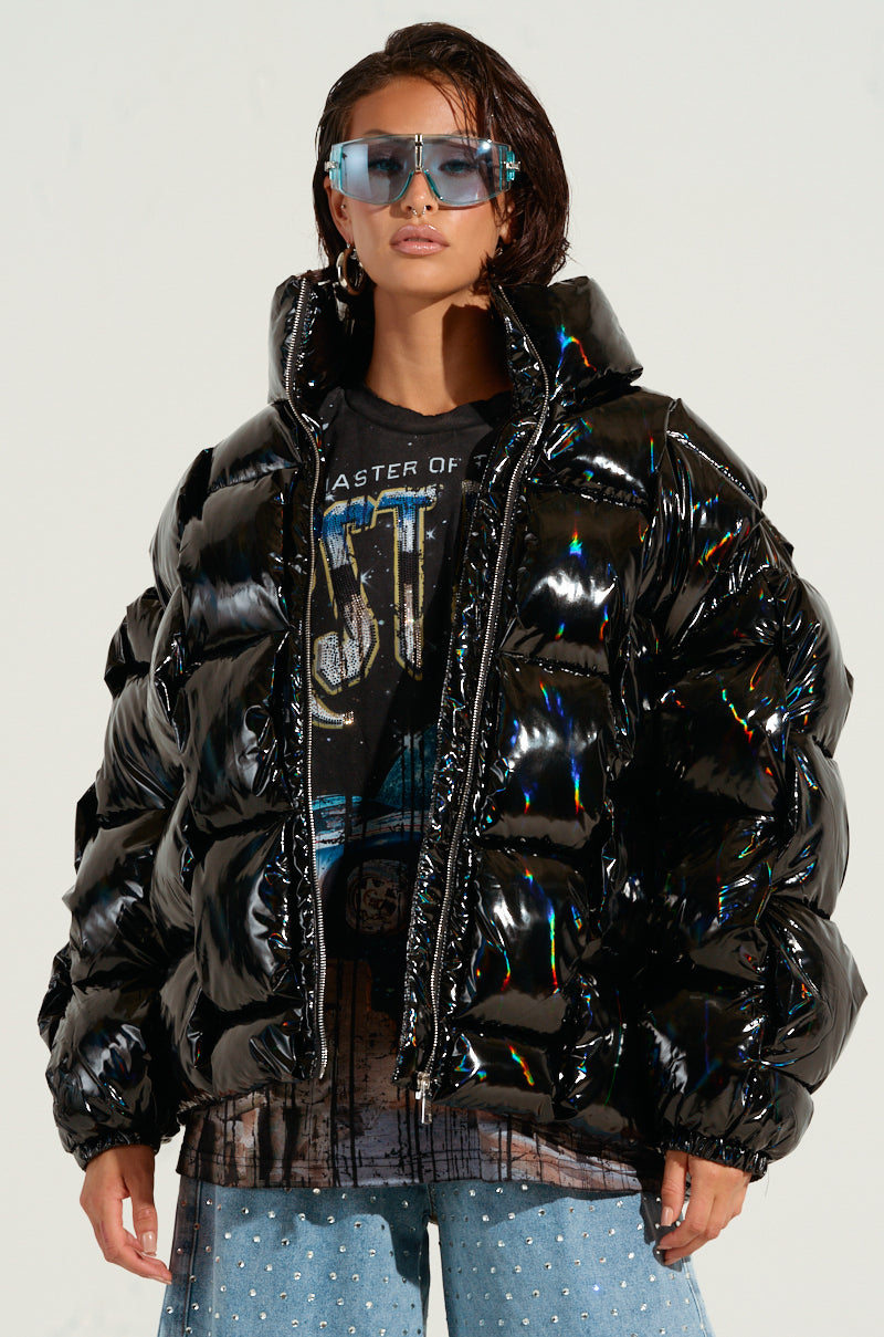 A person is confidently wearing the BOB AND WEAVE OIL SLICK PUFFER, featuring a metallic front zipper, and large sunglasses. They have short hair and are also sporting a graphic T-shirt paired with jeans adorned with jeweled embellishments, set against a plain background.