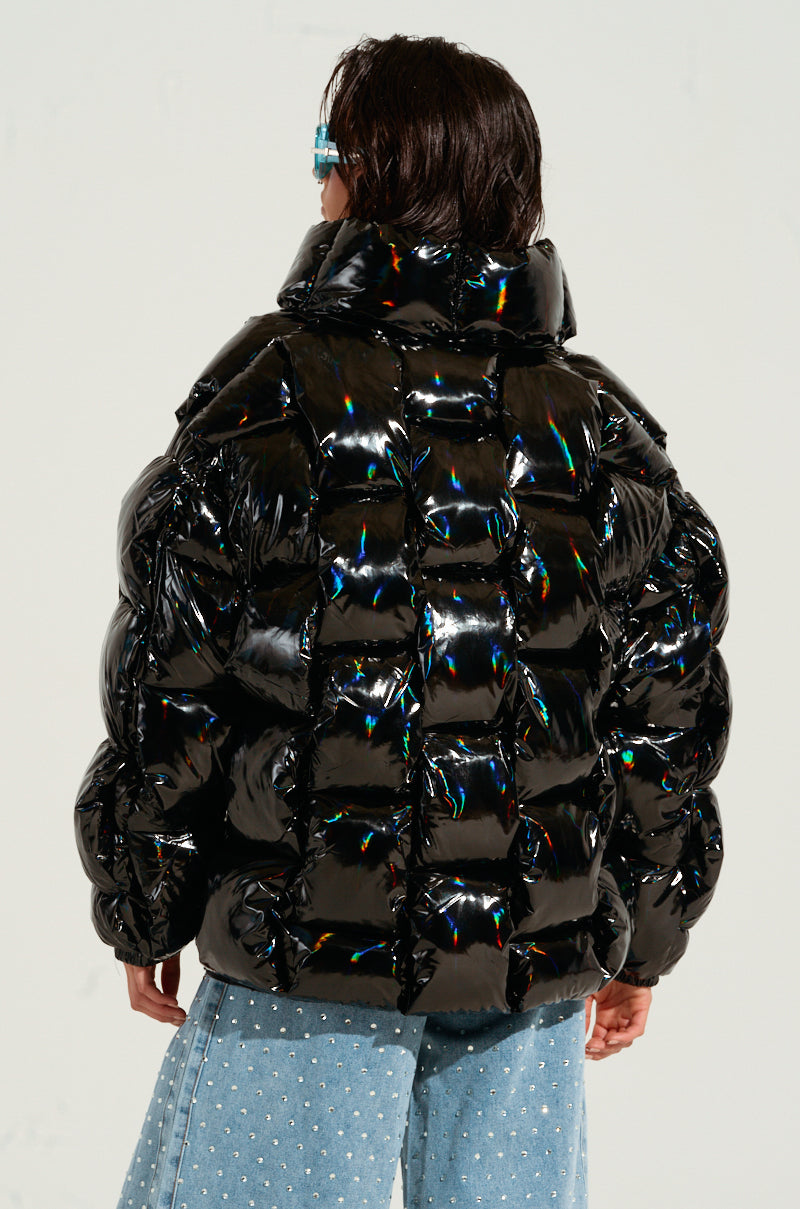 Wearing the BOB AND WEAVE OIL SLICK PUFFER jacket, which features a metallic front zipper, a person with short dark hair stands with their back to the camera. They are also dressed in light blue jeans that have a dotted pattern.