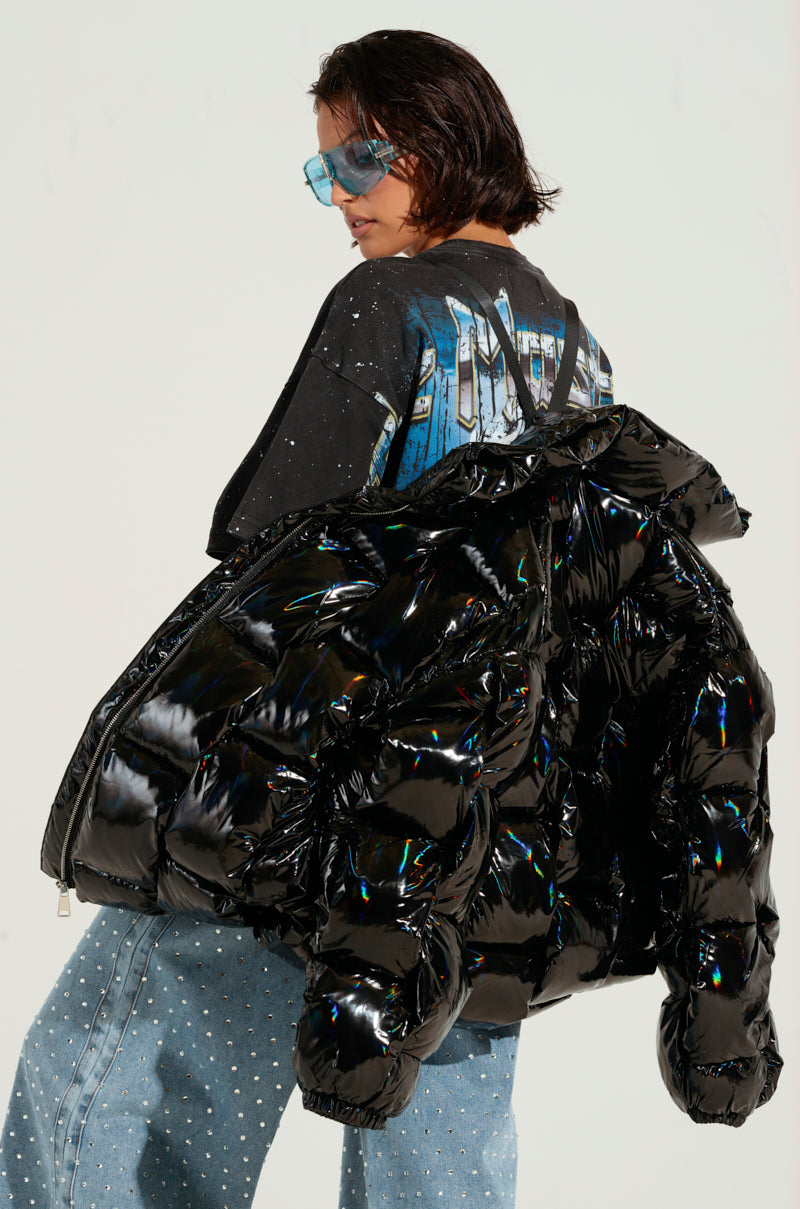 A person with short, dark hair, wearing blue sunglasses, a graphic T-shirt, and oversized blue jeans is seen with the BOB AND WEAVE OIL SLICK PUFFER featuring a metallic front zipper stylishly draped over their shoulder as they face slightly away from the camera.