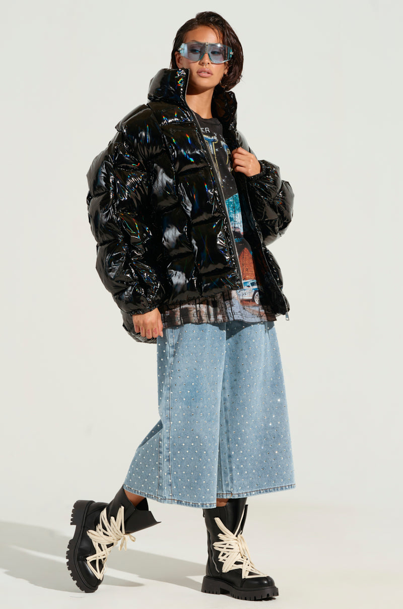A person wearing the BOB AND WEAVE OIL SLICK PUFFER featuring a metallic front zipper, a graphic t-shirt, denim culottes embellished with pearls, black boots with white laces, and sunglasses. They stand confidently against a plain background.