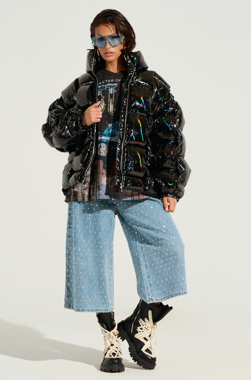 A person wearing the BOB AND WEAVE OIL SLICK PUFFER jacket, a graphic T-shirt, blue wide-leg pants with white dots, chunky boots, and blue sunglasses. They have short dark hair and are posing confidently against a plain background.