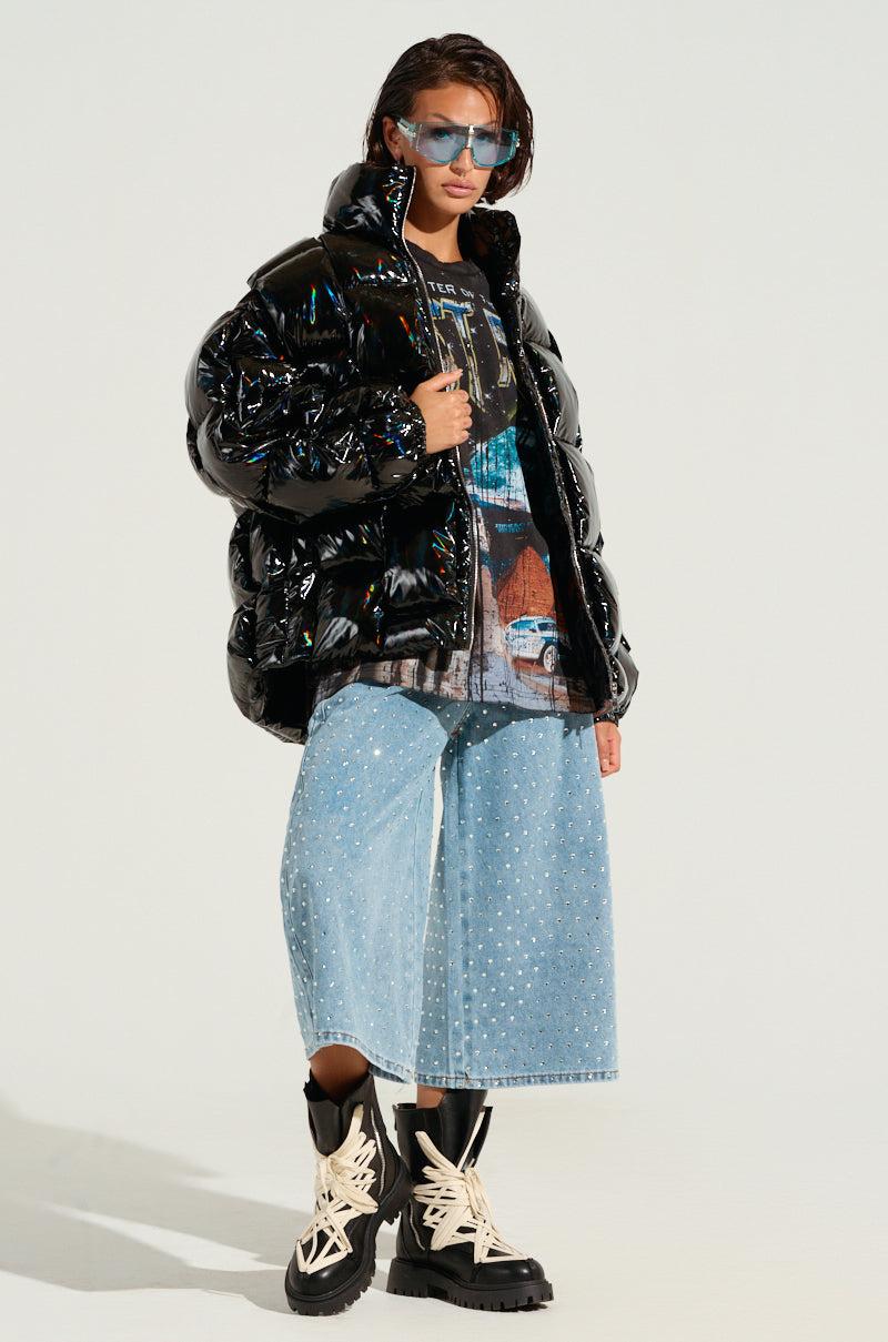 A person dressed in the BOB AND WEAVE OIL SLICK PUFFER featuring a metallic front zipper, paired with a graphic t-shirt, wide-legged light blue jeans, and black boots with white laces. They have short hair and are wearing blue sunglasses while standing against a plain background.