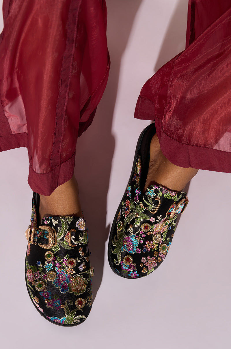 A person wearing transparent, red, loose-fitting pants stands with feet slightly apart, showcasing the AZALEA WANG QUINBY BROCADE CLOG IN BLACK. These black brocade mules feature colorful, intricate floral embroidery and a decorative buckle on each shoe against a plain, light-colored background.
