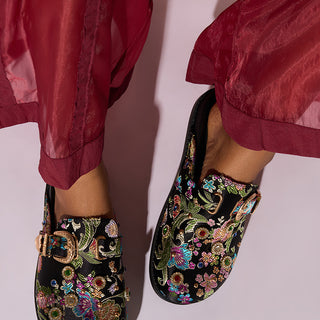 A person wearing transparent, red, loose-fitting pants stands with feet slightly apart, showcasing the AZALEA WANG QUINBY BROCADE CLOG IN BLACK. These black brocade mules feature colorful, intricate floral embroidery and a decorative buckle on each shoe against a plain, light-colored background.