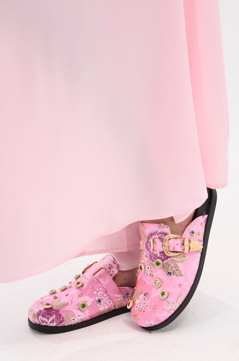 A person in a pale pink, flowing dress stands with feet crossed, showing the AZALEA WANG QUINBY BROCADE CLOG IN PINK—a pink brocade clog adorned with floral embroidery, gemstone accents, sequins, and gold buckles.