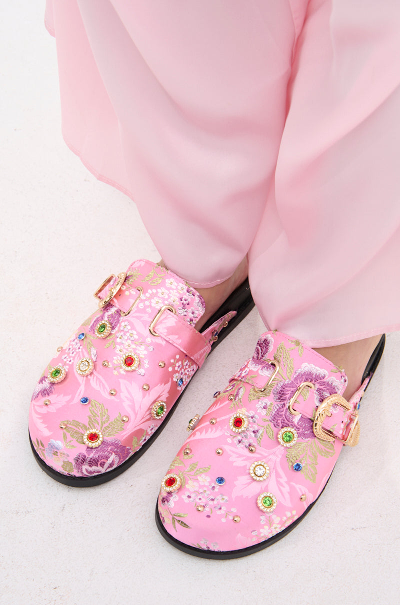 A person wearing light pink pants and the AZALEA WANG QUINBY BROCADE CLOG IN PINK, featuring gemstone accents, embroidered flowers, and gold buckles, stands on a light surface.