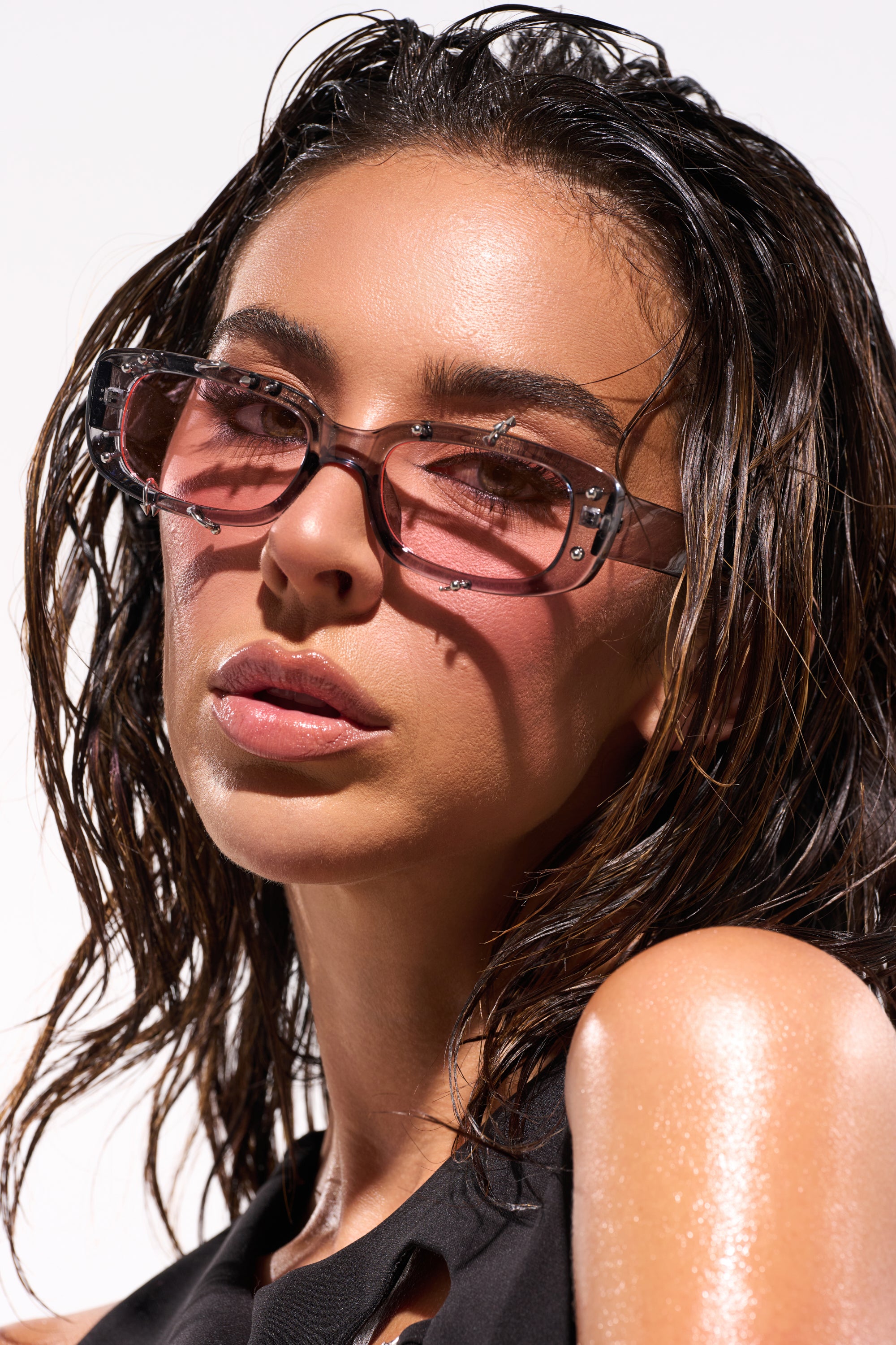 A woman with wet, wavy hair and glowing skin wears PIERCED THRU SUNGLASSES and a black sleeveless top, posing against a bright background. 0
