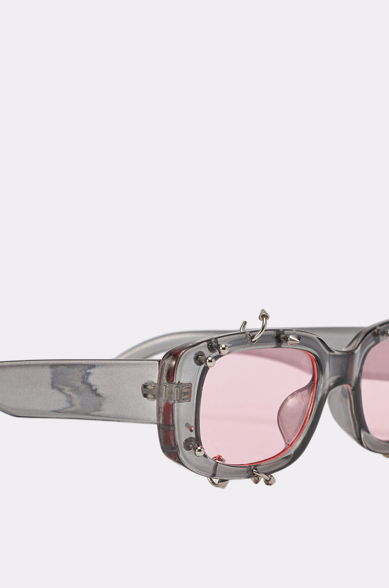 Close-up of PIERCED THRU SUNGLASSES: translucent gray streetwear frames with pink-tinted lenses, detailed with silver screws and bold decorative hardware for an edgy, industrial look. Set against a plain light background. 3
