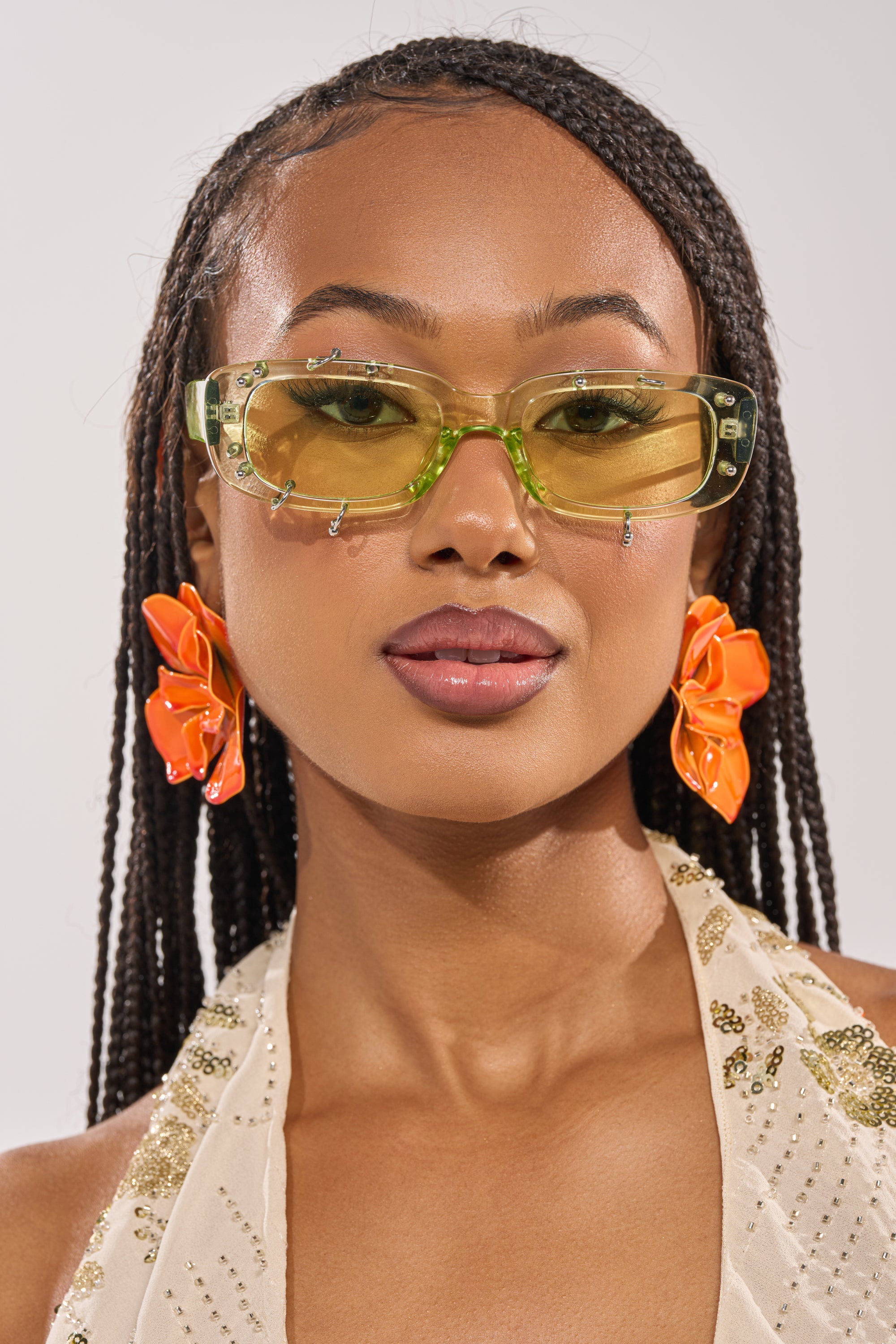 A woman with braided hair confidently wears WITH A TWIST SUNGLASSES with yellow-tinted lenses, large orange flower earrings, and a cream gold sequin halter top against a neutral background. 1