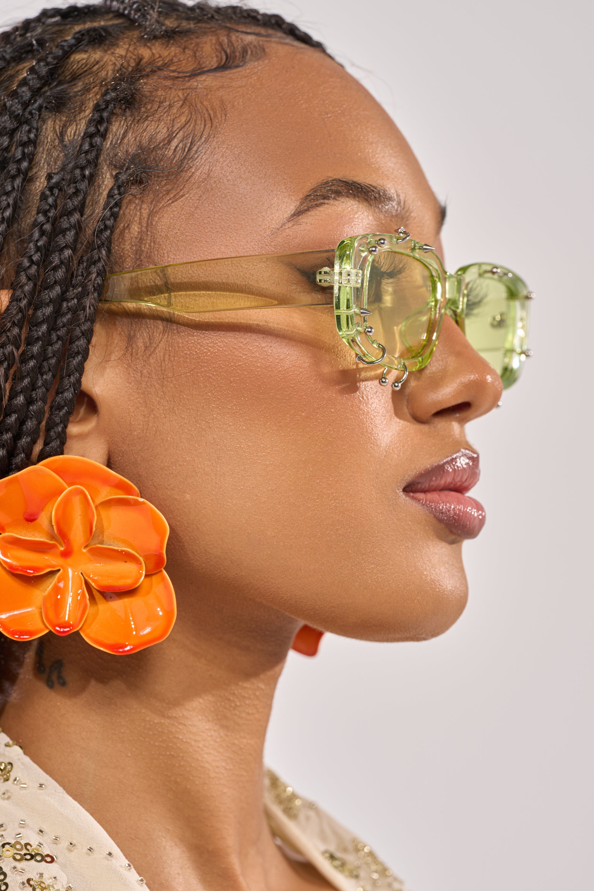 A woman with braided hair wears WITH A TWIST SUNGLASSES in translucent green and bold, orange flower-shaped earrings. Her smooth, glossy skin stands out as she faces right against a plain background. 2