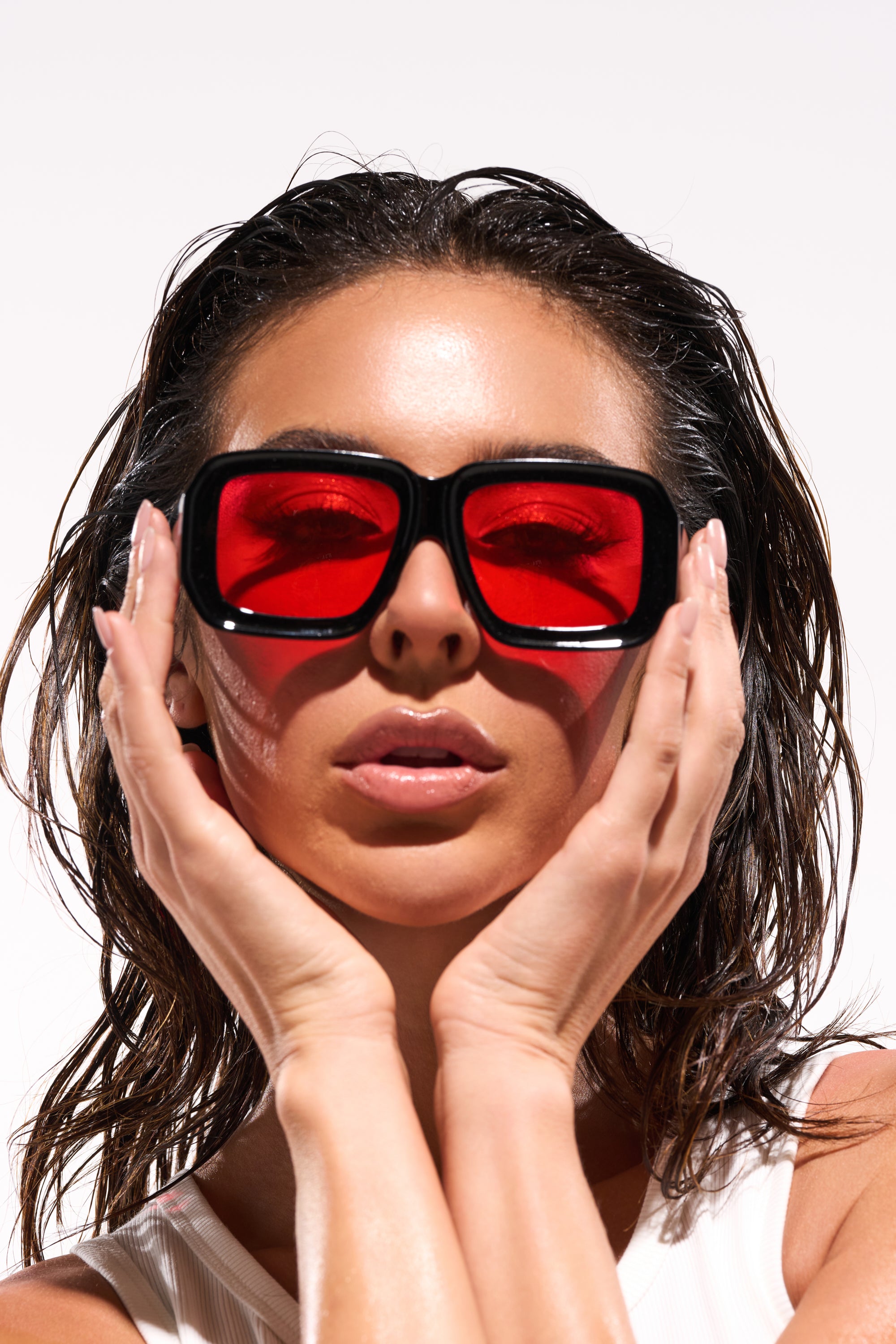 A woman with wet, slicked-back hair poses against a plain white background, touching her face with both hands while wearing RED VISION SUNGLASSES, featuring a bold oversized black and red design. 0