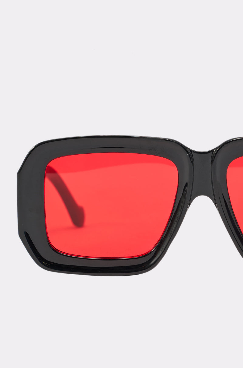 Close-up of one side of RED VISION SUNGLASSES, featuring bold black oversized frames and vibrant red-tinted lenses, set against a plain light gray background. 4
