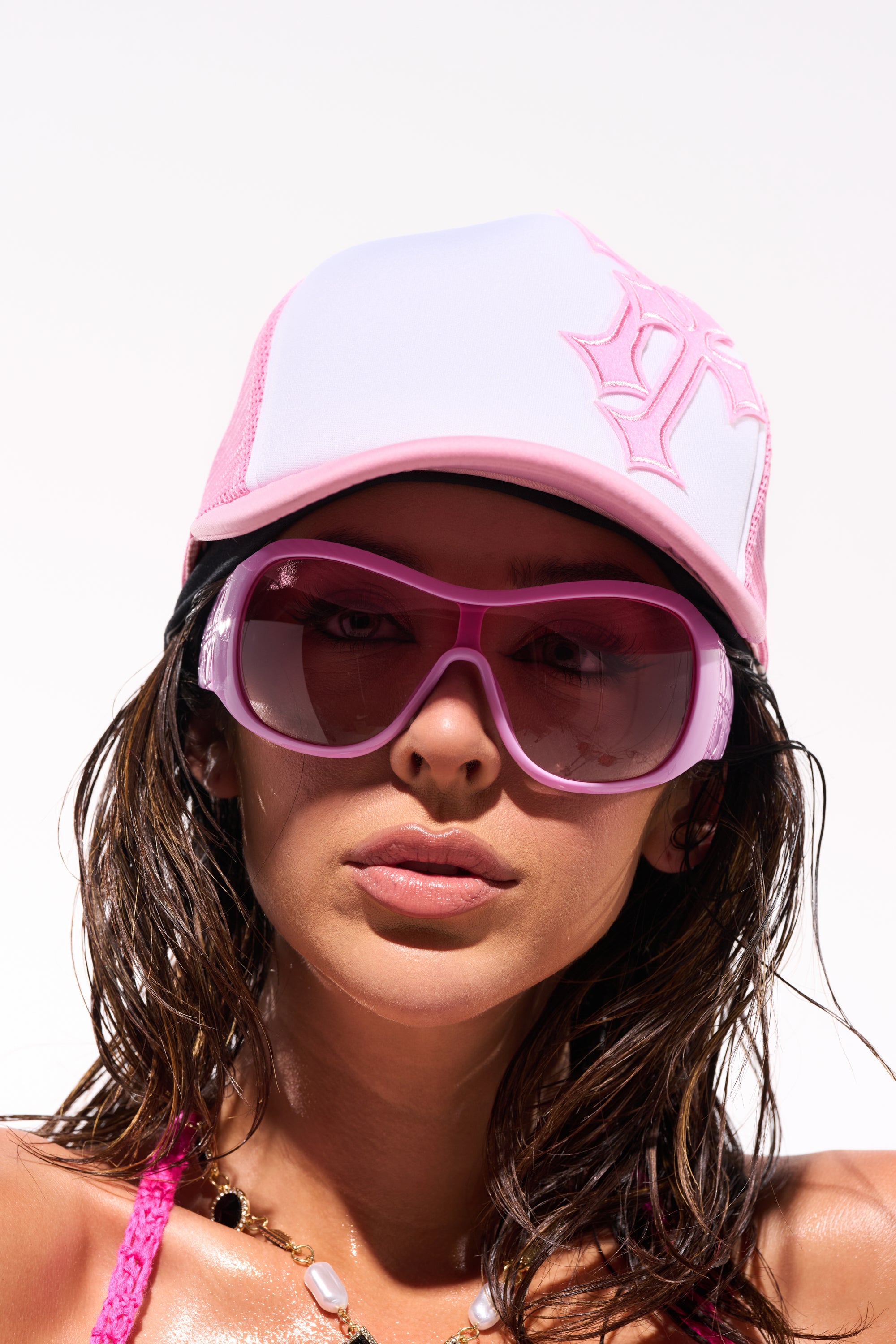 A woman wears BUBBLEGUM SUNGLASSES, a pink and white baseball cap with a large "P," and a pink swimsuit top while posing against a white background. Her hair looks wet, and she completes the look with a beaded necklace. 1