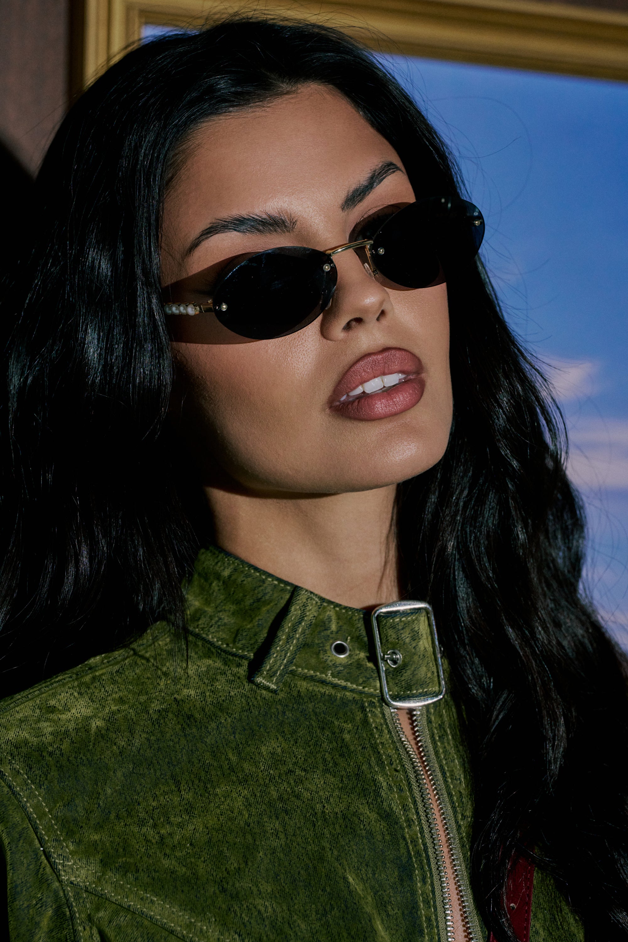 A woman with long dark hair wears SEE YA SUNGLASSES and a green suede jacket, her lips parted confidently. A gold picture frame and blue sky complete the scene in the background. 1